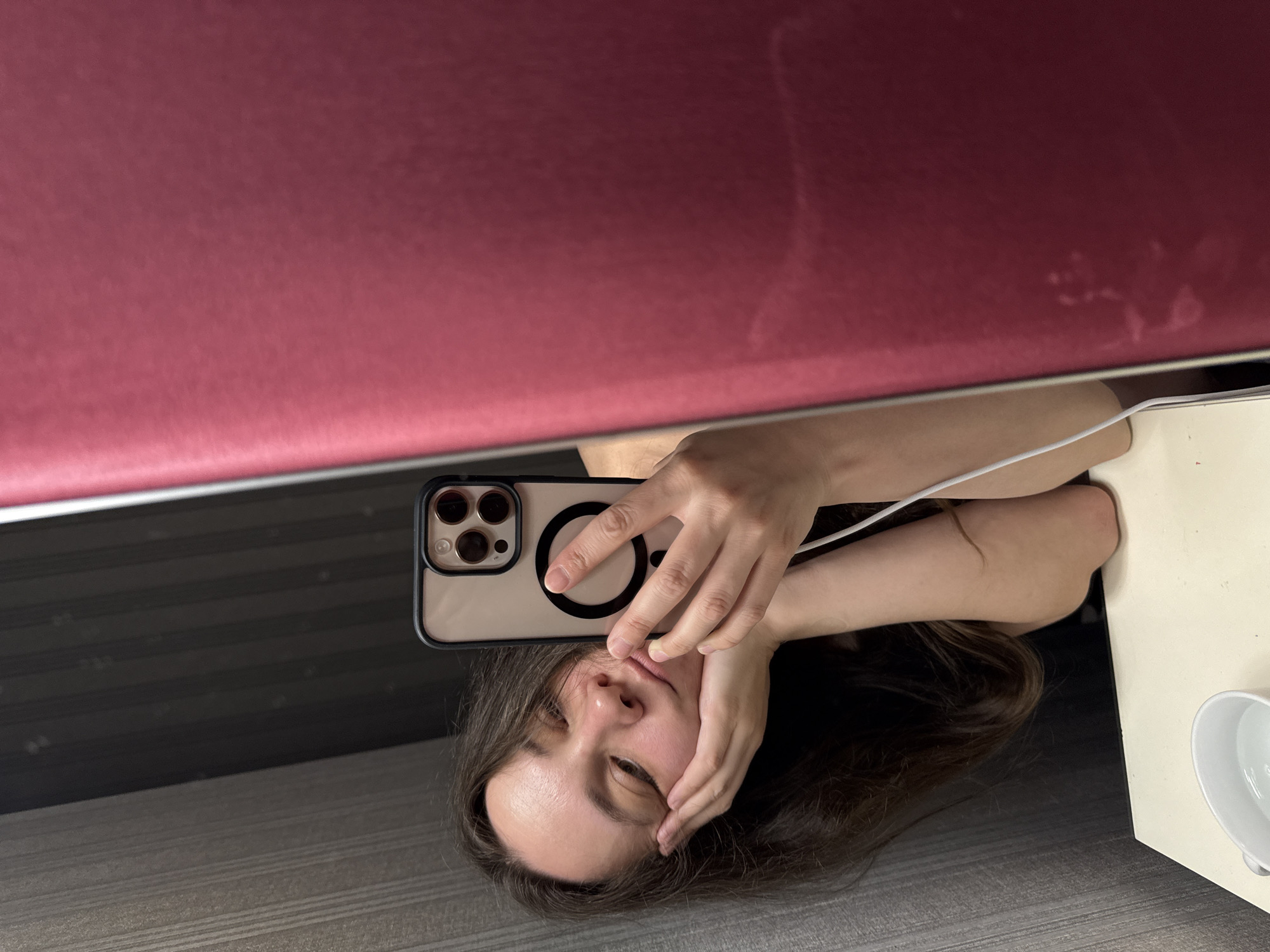 A woman with long brown hair is taking a selfie in a mirror. She is holding her phone, which has a light-colored case with a MagSafe ring, directly above her face. Her head is tilted to the side, resting on one hand. The mirror reflects a burgundy surface. The reflection also shows a table corner with a white surface and a white cup or bowl. The location is not discernible from the image. There is no indication that the photo was taken in Moscow, Russia.