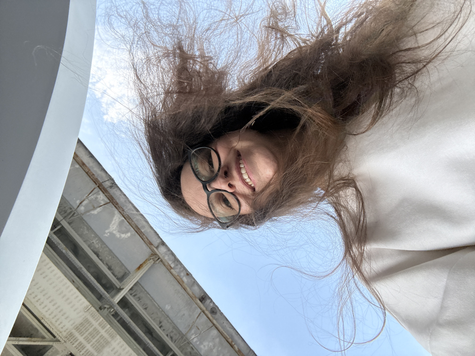 A low-angle shot captures a woman smiling at the camera. She wears glasses and has brown hair that is windswept, obscuring part of the sky behind her. The location appears to be outdoors, adjacent to an industrial or utilitarian structure. Elements of the structure, including a metallic framework and what appears to be a ceiling with gridded patterns, are visible in the lower left of the frame. The background includes a clear blue sky. Given the details, the image is unlikely to contain identifying information to confirm the location in Nizhny Novgorod, Russia.