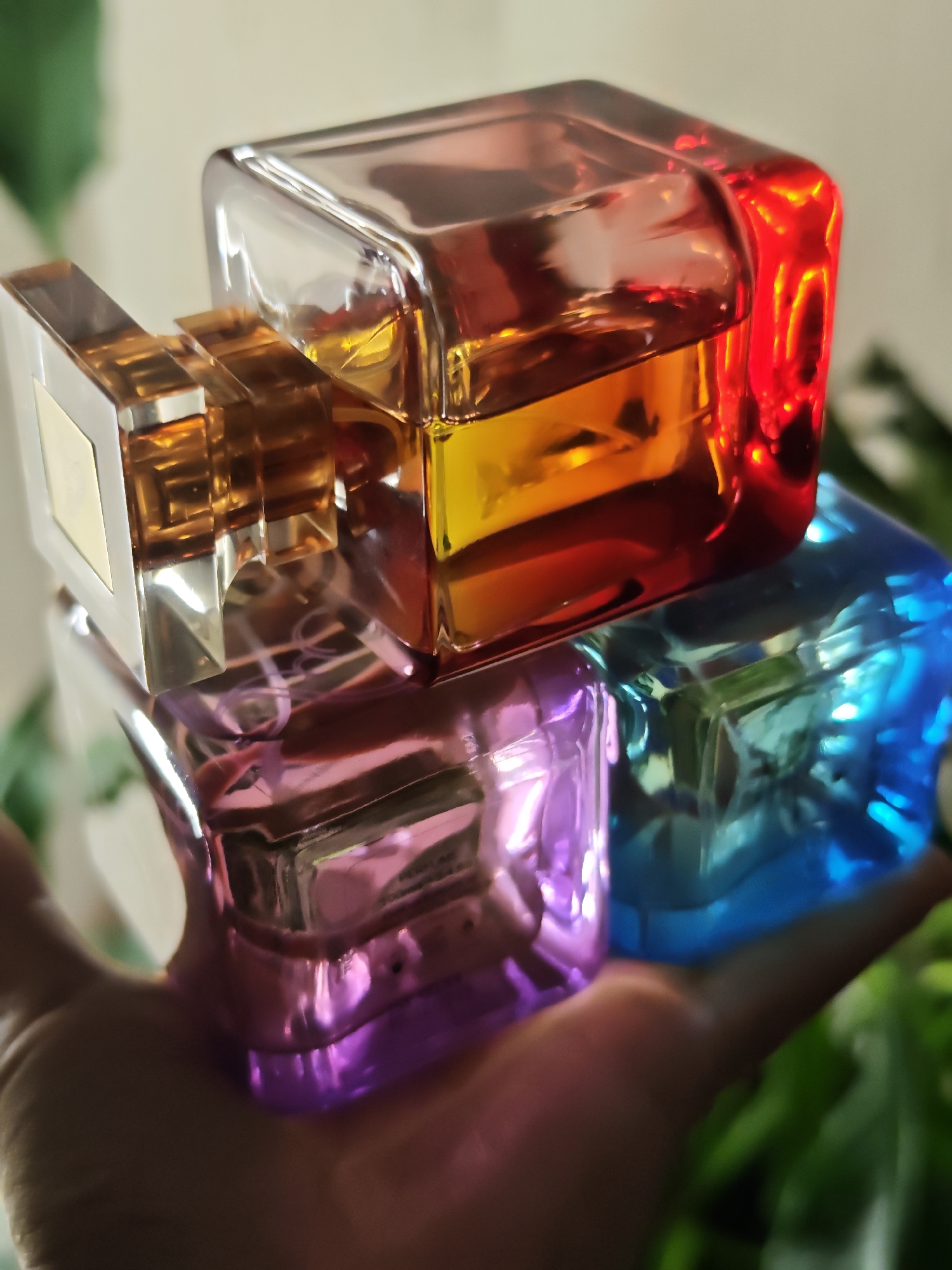 A hand is holding four stacked, colorful perfume bottles. The top bottle has amber and red liquid, the one below it is pink, and the bottom one is blue. The bottles are made of glass and have a faceted design. The background is blurred and appears to be an indoor setting with green foliage visible. There is no indication of the city of Kursk, Russia.