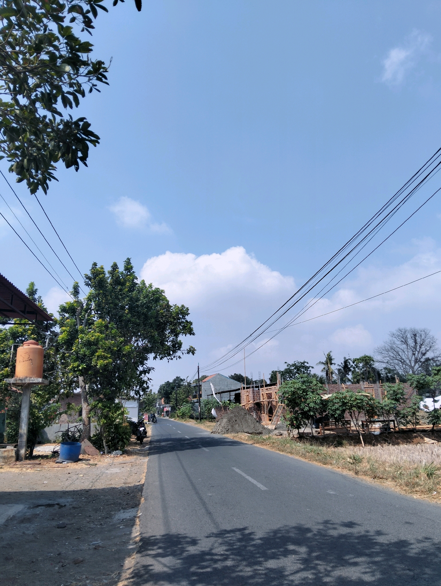 The image depicts a rural road in Banjarparakan, Indonesia, under a bright blue sky with scattered white clouds. The road, paved with asphalt, runs diagonally from the lower right to the upper left of the frame, casting shadows from nearby trees onto its surface. On the left side of the road, a large, leafy tree stands prominently, with a water tank on a concrete pillar situated in front of it. A blue plastic barrel is also visible near the base of the tree.
Further down the road, on the left, is a building with a corrugated metal roof. Beyond that, the road curves and is lined with more trees and houses. On the right side of the road, construction is underway on what appears to be a building, with scaffolding and piles of construction material. Several smaller trees and dry vegetation border the construction site. In the distance, a few motorcycles are seen on the road, suggesting some activity. The overall scene conveys a sense of everyday life in a developing rural area.
