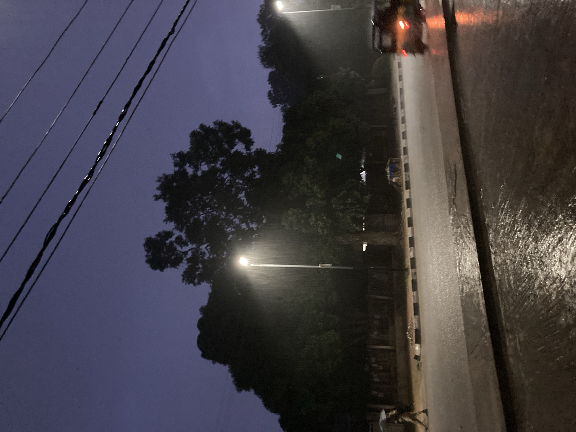 The image depicts a street scene in Jos, Nigeria, during a heavy downpour. The sky is a deep twilight blue, indicating it is either early evening or late afternoon. Power lines are visible crisscrossing the upper part of the frame. A solitary street lamp is illuminated, casting a bright, upward-pointing beam that illuminates the falling rain, creating a misty effect. Large, dark trees with dense foliage form a silhouette against the sky, suggesting a roadside park or residential area.
On the wet asphalt road, which reflects the ambient light and the glow from the streetlamp, a person is walking under an umbrella, their figure partially obscured by the rain. Further down the road, a three-wheeled vehicle, possibly a tricycle or auto-rickshaw, is visible with its taillights illuminated, indicating it is moving. The road surface is slick and appears to have standing water in places, consistent with heavy rainfall. The overall atmosphere is somber and atmospheric, conveying the mood of a rainy evening in the city.