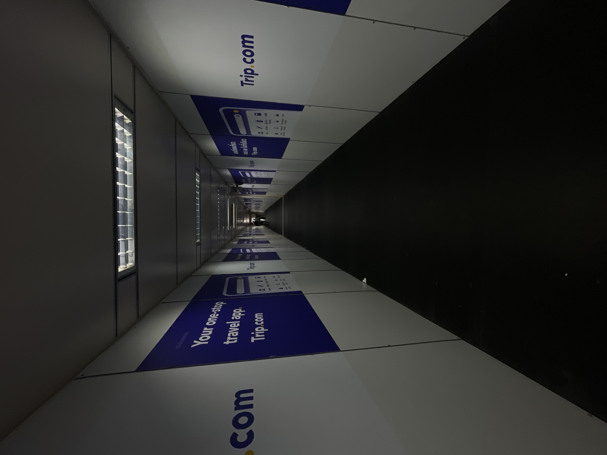 An interior shot shows a dimly lit, long corridor with white walls displaying advertisements for Trip.com. The corridor appears to be under construction or renovation, with the white panels serving as temporary covering. Fluorescent lights are visible in the ceiling, providing minimal illumination. The floor is covered with a dark material, possibly carpet or rubber matting. The perspective is from the beginning of the corridor, looking towards a faint light at the far end, where a solitary figure is dimly visible. The advertisements are consistent on both sides of the corridor, featuring the Trip.com logo and promotional text in blue and white. The overall atmosphere is utilitarian and somewhat sterile.