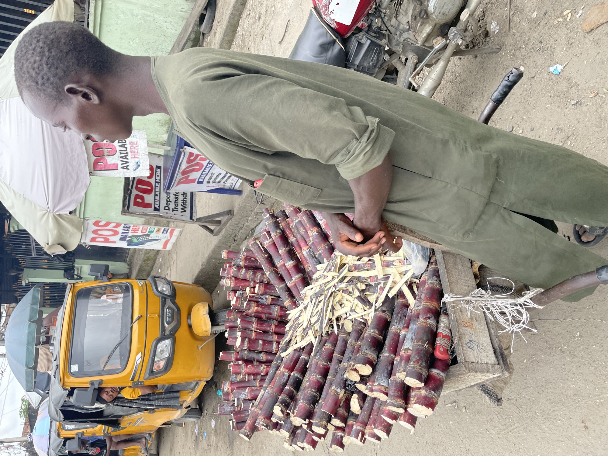 In Ado, Nigeria, a vendor stands next to a cart piled high with sugarcane stalks. The sugarcane, a deep reddish-purple, is neatly arranged in the wooden cart, with some stalks appearing to have been partially peeled. To the left of the cart, a yellow auto-rickshaw, also known as a Keke Napep, is parked. A man is visible sleeping in the driver's seat of the auto-rickshaw. Behind the vendor and the cart, a motorcycle is parked. Several signs with the word "POS" are visible in the background, indicating point-of-sale services are available. The ground is made of a rough, sandy concrete. The overall scene suggests a street-side market or commercial area.