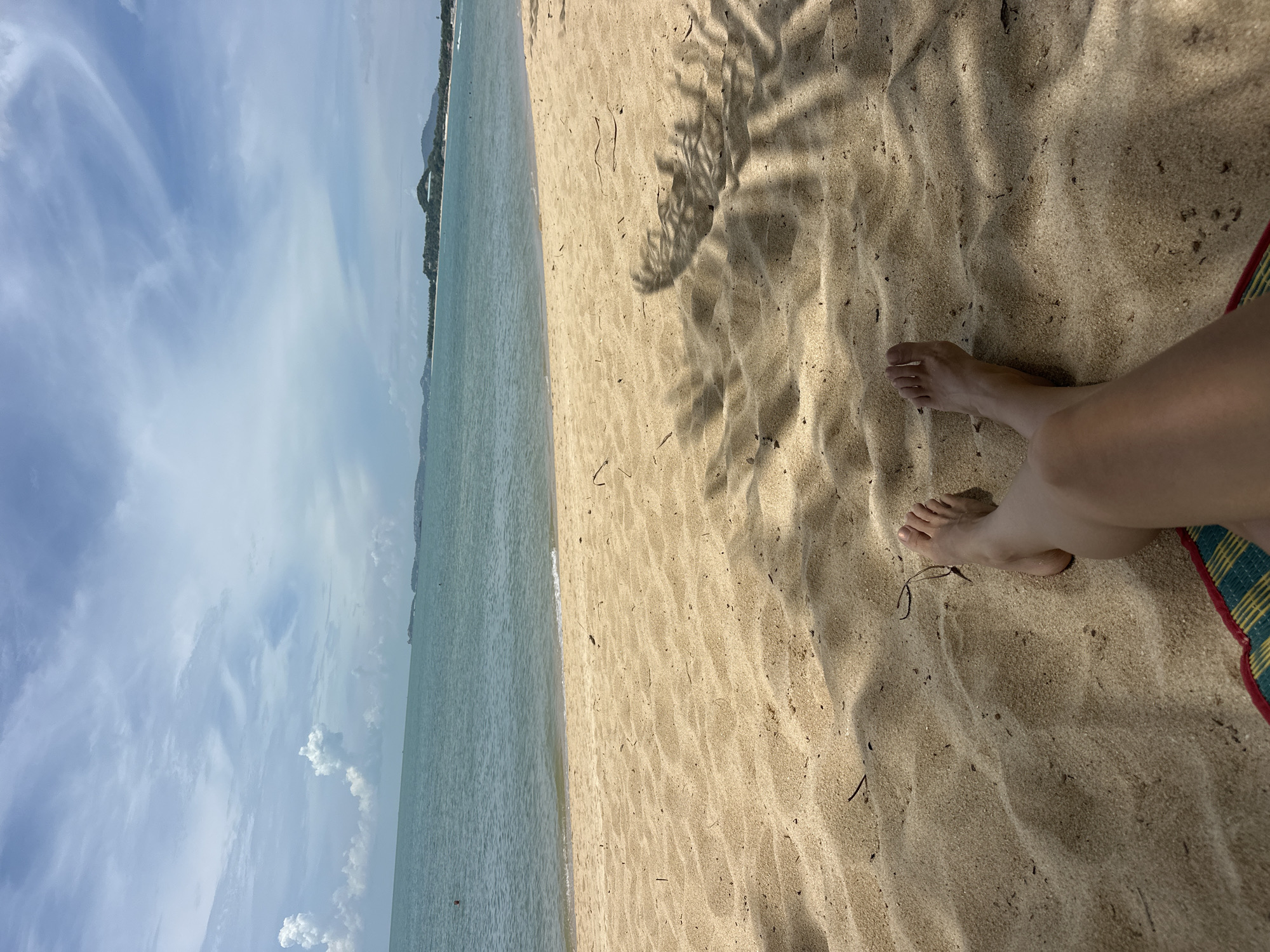 The image depicts a serene beach scene in Baan Mae Nam, Thailand. The foreground is dominated by golden sand, casting shadows from palm fronds overhead. In the center of the frame, a person's bare legs and feet are visible, suggesting a relaxed posture, perhaps lounging on a mat, indicated by the colorful fabric at the bottom edge. Beyond the sand, the calm turquoise water of the sea stretches to the horizon, where a faint coastline with lush green hills and scattered buildings can be observed. The sky is a vibrant blue, adorned with fluffy white clouds, adding to the tranquil atmosphere. The overall impression is one of a peaceful and idyllic day at the beach.