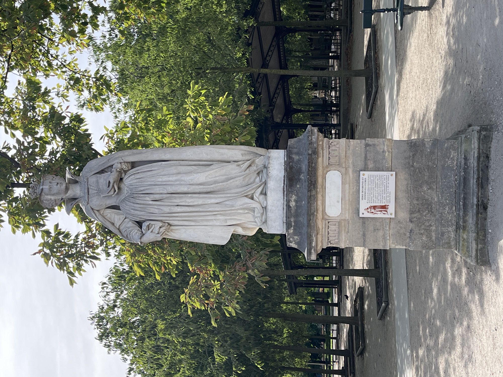 The image shows a statue of a queen in a public park. The statue is made of white marble and depicts a woman wearing a crown, a long dress, and a cape. She is holding a book in her left hand and her right hand is resting on her hip. The statue is on a pedestal that has a plaque with French text and an illustration. The park is filled with trees and has pathways, benches, and covered seating areas. The sky is overcast, and the lighting suggests it is daytime. The overall scene is tranquil and orderly. The statue is located in the Luxembourg Gardens in Paris, France.