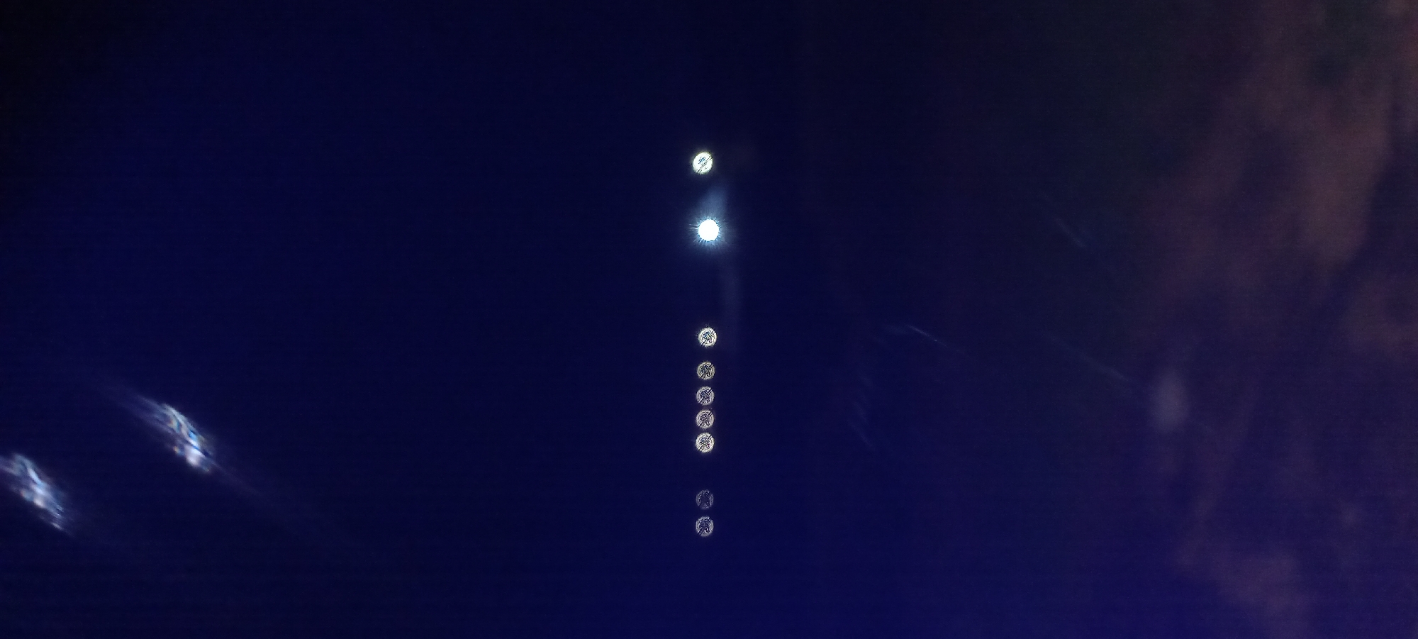 The image depicts a dark, night sky with several bokeh effects, suggesting artificial lights that are out of focus. Two prominent, elongated blurry lights with a prismatic effect are visible in the upper portion of the frame. Below these, a line of seven circular bokeh lights is present, with one significantly brighter than the others, indicating a strong light source. The lower part of the image shows a dimly lit, cloudy or hazy area, possibly representing the lower atmosphere or ground cover. The overall scene is devoid of identifiable human figures or specific landmarks that would pinpoint its location within Kwang, Nigeria. The composition suggests an observation of lights from a distance against a dark backdrop.