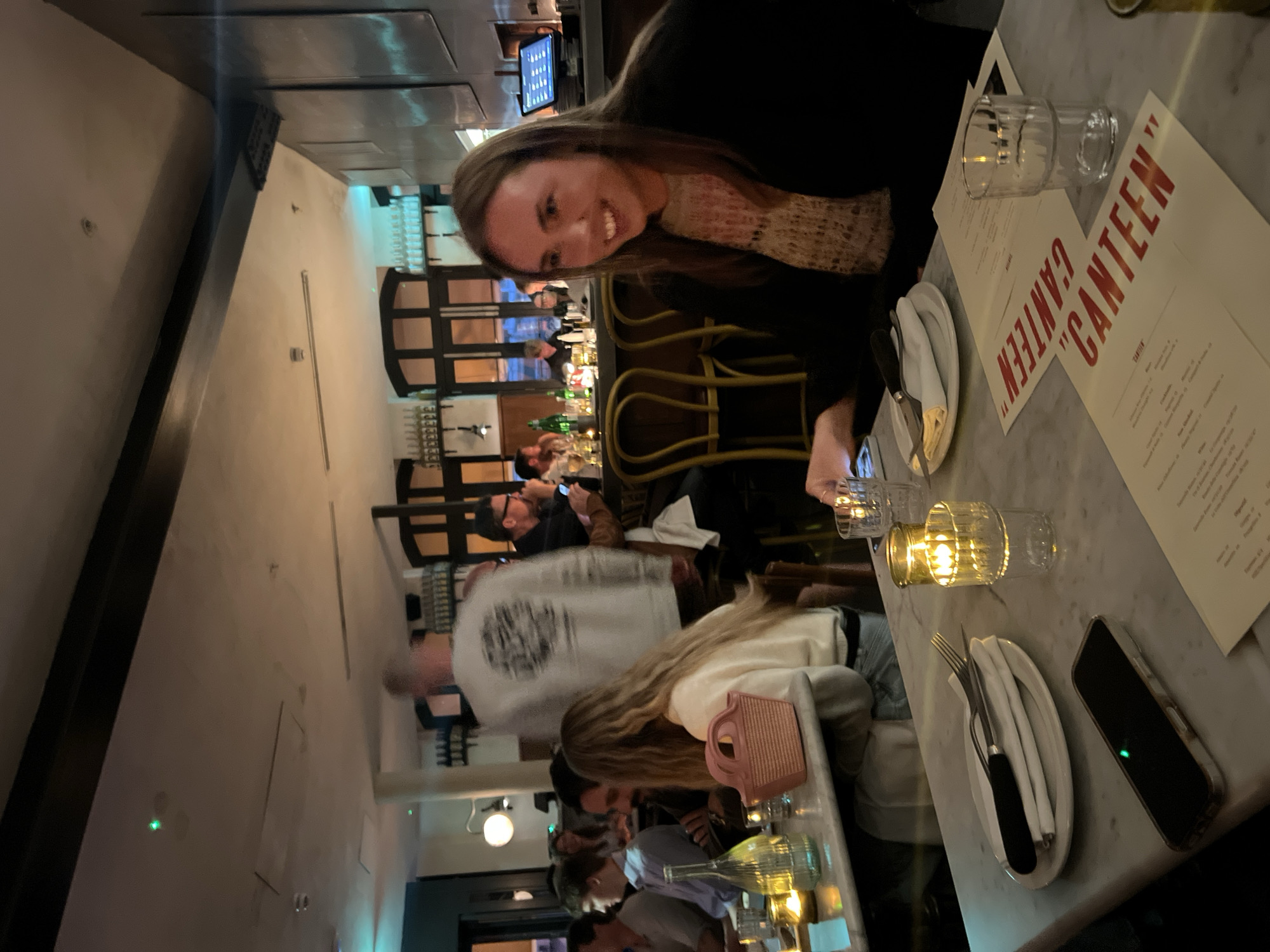 The image captures a dimly lit restaurant interior. A woman smiles at the camera from a table with marble finish, on which a menu card with "CANTEEN" emblazoned in red letters, tableware, a glass, and a phone are placed. A candle holder emits a soft glow. The background shows a busy restaurant scene with people seated at the bar and tables, some standing and walking. A man in a grey hooded sweater is partially blurred, suggesting movement. The restaurant appears to be in London, United Kingdom, based on the restaurant name and typical London restaurant interior designs.