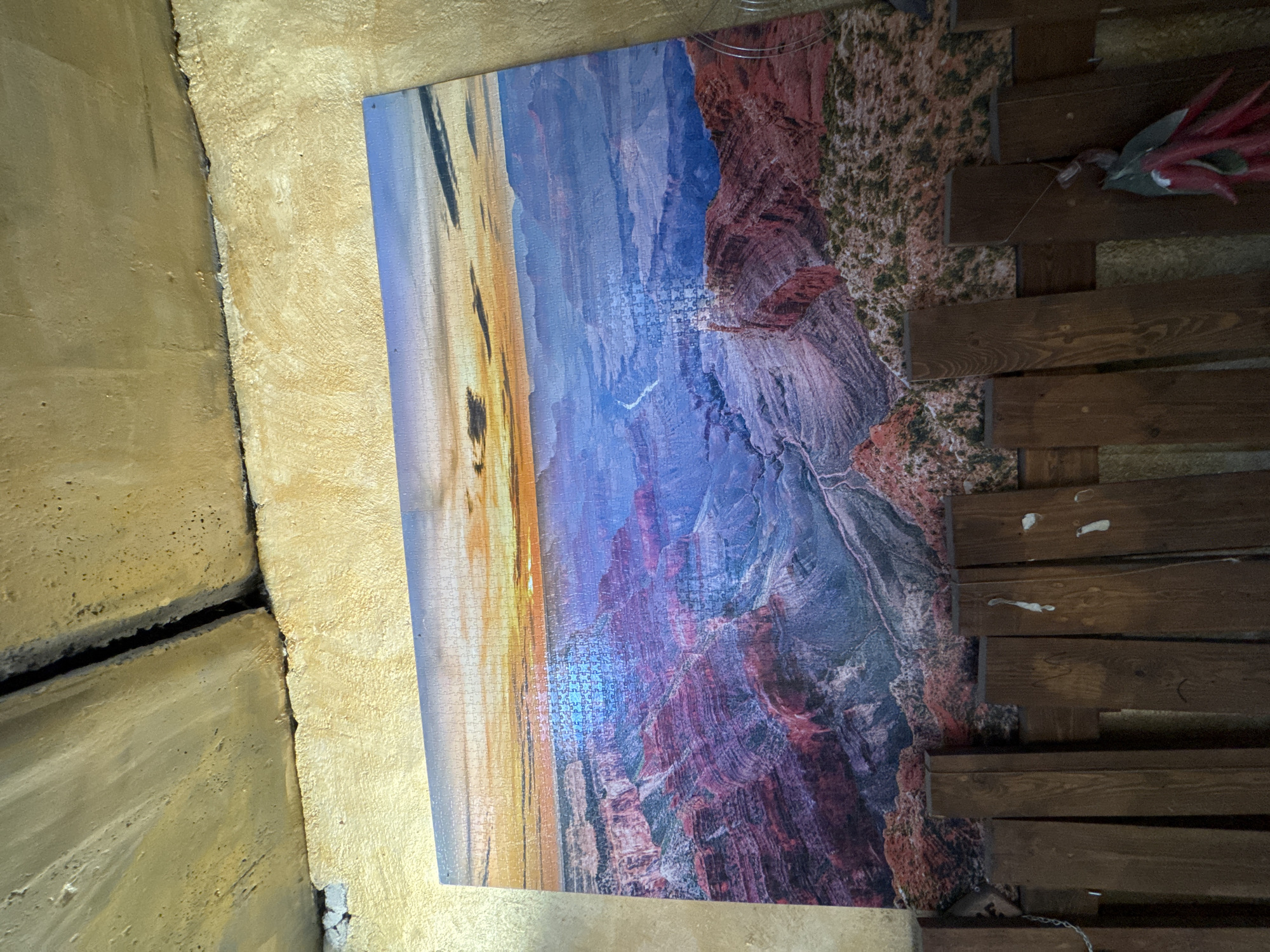 This image captures a large jigsaw puzzle depicting a scenic landscape, likely the Grand Canyon, displayed on a wall in an indoor setting in Yerevan, Armenia. The puzzle, viewed as if horizontally oriented, shows a vast canyon with layers of rock formations. The left side of the puzzle features warm hues of orange and yellow, characteristic of sunrise or sunset, while the right side transitions into cooler blue and purple tones, illustrating the depth and shadows of the canyon. A distinct area in the mid-right section of the puzzle appears incomplete, revealing a grid of missing pieces.
To the left of the puzzle, a light beige or cream-colored wall with a textured, stucco-like finish is visible. A prominent, dark, uneven crack runs vertically through this wall. On the right side of the puzzle, a section of dark brown wooden planks or slats is arranged horizontally, forming part of the wall. These planks display natural wood grain and some small white markings. A decorative element with red, leaf-like shapes is visible towards the upper right, seemingly attached to the wooden structure with thin wires.
The scene is static, featuring no people or active event, focusing solely on the displayed puzzle and its immediate surroundings. The lighting suggests an indoor environment. No discernible text is visible anywhere in the image.