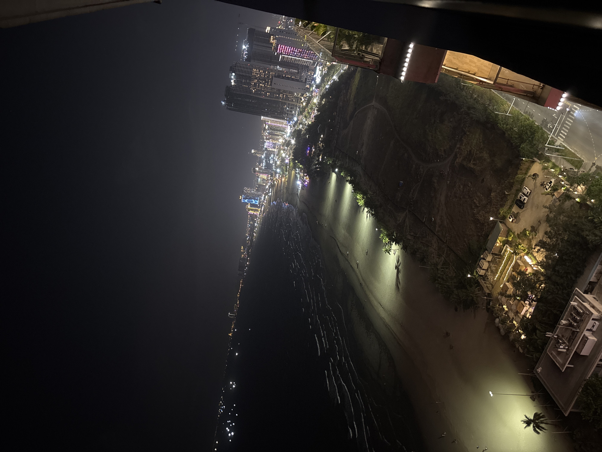 This high-angle, nighttime shot captures the coastal cityscape of Đà Nẵng, Vietnam. The image is dominated by the dark expanse of the ocean on the left, with gentle waves creating luminous white lines as they break on the shore. The beach itself is a strip of sand, sparsely populated with a few faint human figures and illuminated by scattered artificial lights casting long shadows from palm trees.
Further inland, the city skyline glows with a multitude of lights from numerous high-rise buildings, creating a vibrant urban tapestry against the dark sky. The buildings vary in height and architectural style, with some featuring colorful illuminated facades, suggesting a lively commercial and residential area. A coastal road is visible, lined with trees and punctuated by streetlights and crosswalks, with several parked cars visible on its side. In the foreground, a portion of a building or balcony railing, possibly from the vantage point of the photographer, is visible on the right, featuring a series of bright lights. The overall impression is a dynamic and illuminated coastal city at night, showcasing the blend of natural and urban elements.