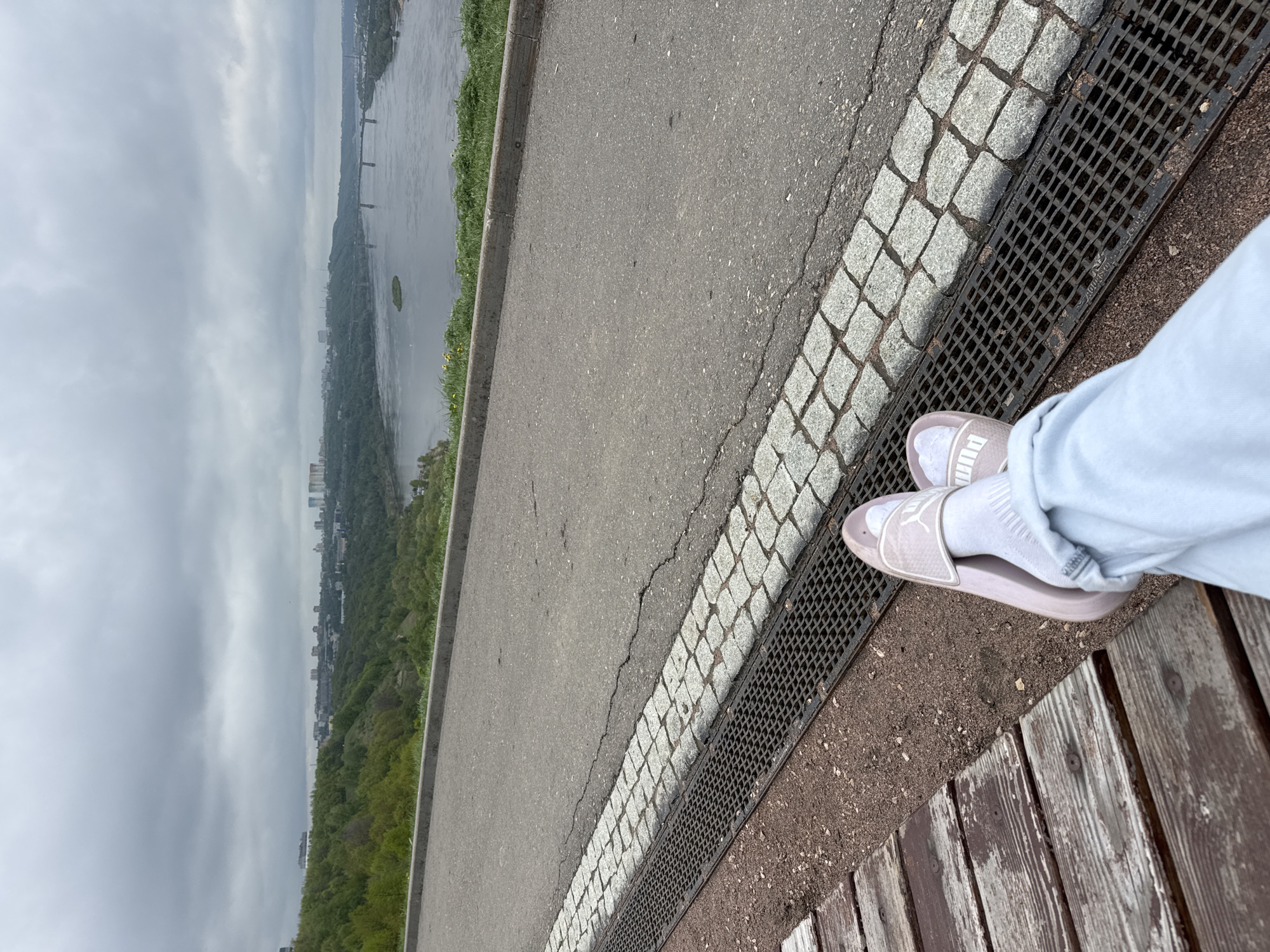 The image shows a view from a high point overlooking the city of Nizhny Novgorod, Russia. The sky is overcast, and the Volga River is visible in the distance with a bridge spanning its width. The foreground features a section of a paved road bordered by cobblestones and a metal grate. A person is seated on a wooden bench with their feet visible, wearing white socks and pink Puma slides. The bench is adjacent to the metal grate. The overall scene suggests a viewing area or a public space along an elevated walkway or park area.