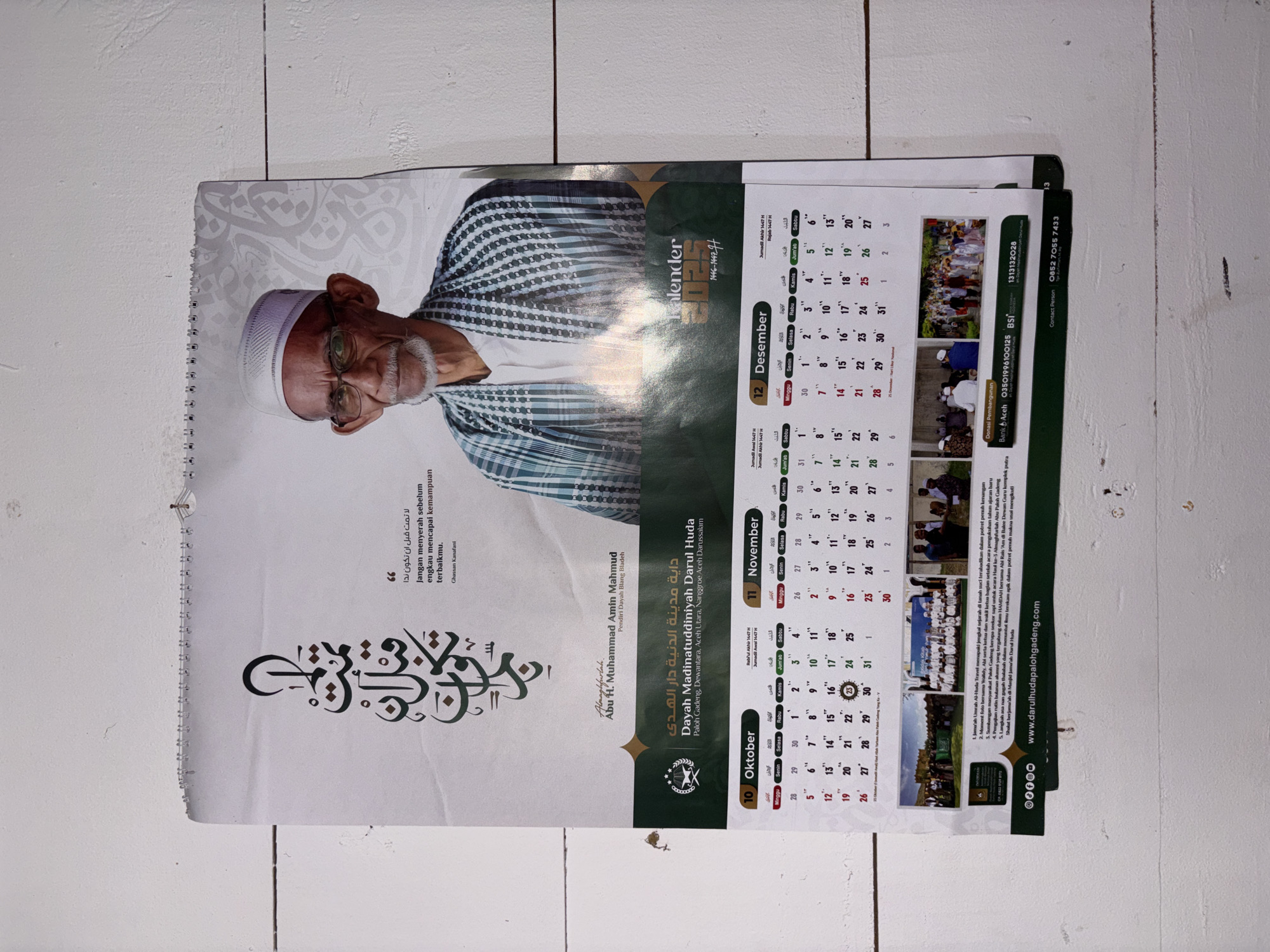 This is a photograph of a wall calendar for the year 2023. The calendar features a portrait of an elderly man with glasses and a white cap, wearing a checkered shirt. He is positioned on the upper right side of the calendar. To his left, there is Arabic calligraphy and a quote in both Arabic and Indonesian: "Jangan menyerah sebelum engkau mencapai kemampuan terbaikmu" (Never give up before you reach your best ability) by Ghassan Kanafani. Below the portrait, the calendar displays information about "Dayah Madinatuludiniyah Darul Huda" located in Paloh Gadeng, Dewantara, Aceh Utara, Nanggroe Aceh Darussalam, Indonesia. The calendar shows the months of October, November, and December, with their corresponding dates and days of the week in both Gregorian and Hijri calendars. There are also photographs of events and a mosque at the bottom of the calendar. The calendar is hanging on a white wall. The provided crops show details of the man's face, the calligraphy, the calendar's grid, and the photographs at the bottom. The location mentioned on the calendar, Paloh Gadeng, Dewantara, Aceh Utara, Nanggroe Aceh Darussalam, can be considered the specific locale within Indonesia.