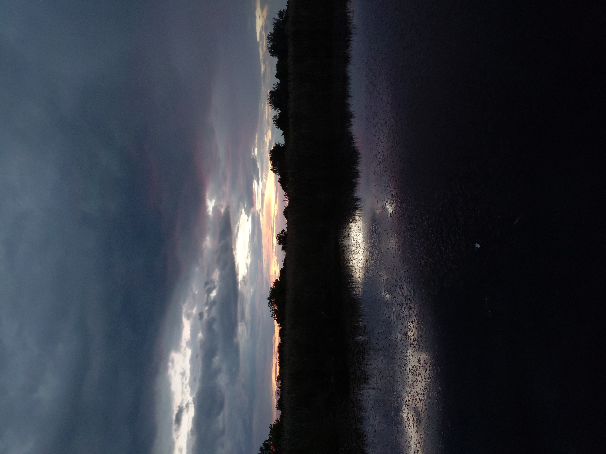 A twilight scene captures a tranquil body of water in the foreground, reflecting the dramatic, cloud-filled sky. A dark, silhouetted line of trees and reeds forms a dense border along the far bank, separating the water from the horizon. The sky is a mosaic of dark, heavy clouds interspersed with lighter patches where the last rays of the sun break through, casting an orange and pink glow. The water's surface is disturbed by ripples, creating a shimmering effect that mirrors the celestial display above. The overall impression is one of natural serenity at the close of day. This scene is located in the village of Ustyvytsia, Ukraine.