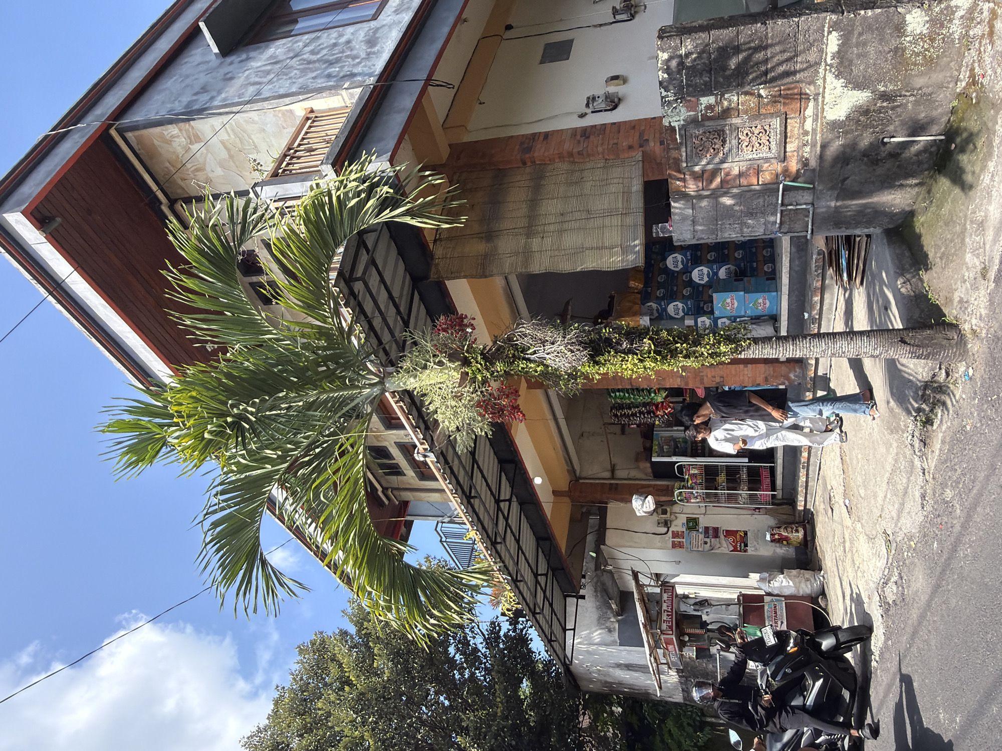 This is an outdoor, eye-level, medium shot of a street scene in Ubud, Indonesia, on a clear, sunny day. The focal point is a building that appears to be a convenience store or small market. The building is multi-story with a grey exterior and wooden trim under the roof. There is a prominent palm tree with green fronds in the foreground, obscuring some of the building's facade.
In front of the building, a man wearing a black jacket and helmet is seated on a black motorcycle, facing left. Next to him, to the right, are two men. One is wearing a white shirt and pants, and the other is in a black t-shirt and blue jeans. They appear to be conversing.
The store entrance is open, revealing shelves stocked with goods. Bottles of "AQUA" and "Le Minerale" water are visible on display. A woven bamboo blind hangs over a portion of the entrance. There are gas pumps labeled "PERTAMINI" to the left of the store. The ground in front of the store is concrete and shows some signs of wear and pooling water. A low stone wall with decorative carvings borders the right side of the building's foundation. Lush green trees are visible in the background. The sky is bright blue with scattered white clouds.