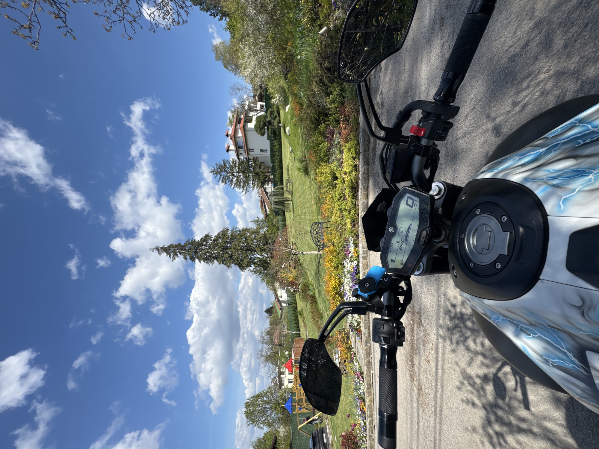 The image shows the point of view from the handlebars of a motorcycle, parked on a paved road. In the background is a residential area with houses, a well-manicured lawn, flowerbeds, and trees under a blue sky with scattered clouds. The motorcycle has a black and gray design with lightning bolt accents. The digital display on the motorcycle shows a reading of "406" and "946." It is not possible to confirm the location as Sofia, Bulgaria based on the image alone.