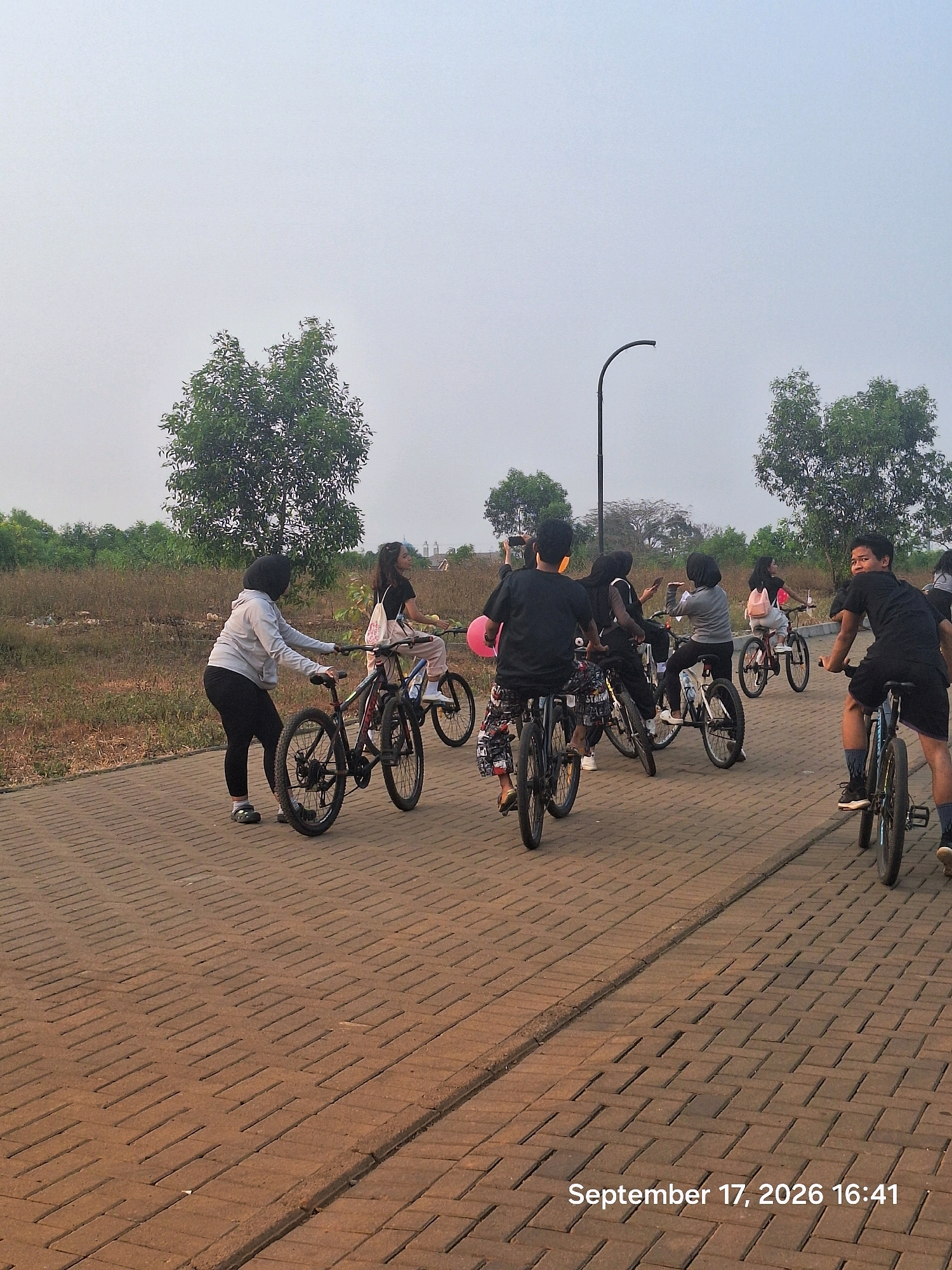 In Depok, Indonesia, on September 17, 2026, at 16:41, a group of young people are cycling on a brick-paved path. The scene is outdoors with a hazy sky and sparse vegetation in the background, including trees and dry grass. A lamppost is visible in the middle ground. The individuals are diverse in gender and are dressed casually. Some are riding bicycles, while others are walking alongside them. One person in the foreground is looking towards the camera and smiling. The overall atmosphere appears to be a recreational outing.
