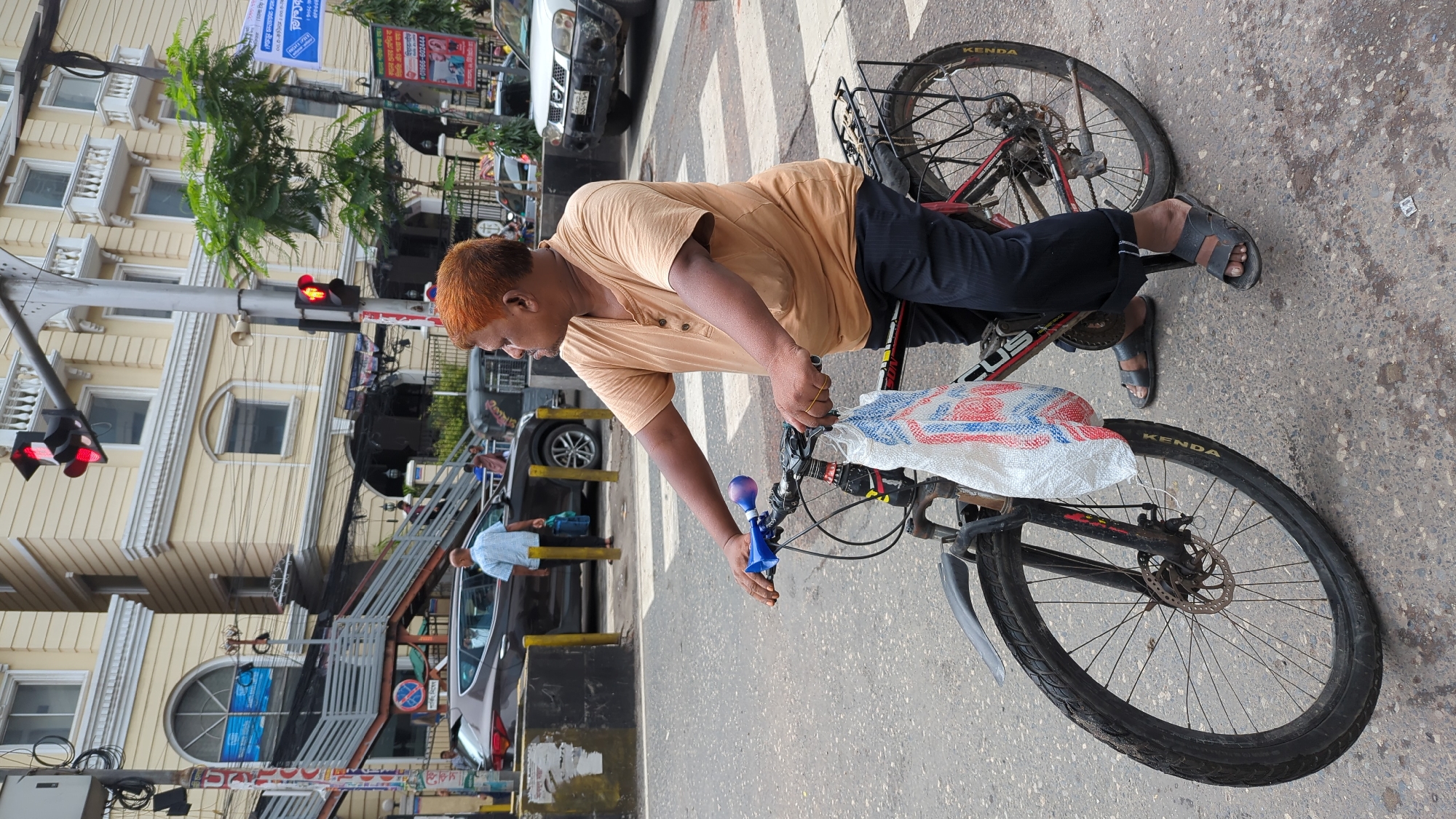 The image depicts a man with reddish-brown hair riding a bicycle at a street intersection in Dhaka, Bangladesh. He is wearing a light brown, short-sleeved shirt with buttons and dark trousers. The bicycle has a white bag with red and blue markings attached to the front. The man is looking to his right, and his hands are on the handlebars. He is wearing black sandals.
In the background, there is a multi-story building with cream-colored walls and white decorative trim. Traffic lights are visible, with one showing red and another showing yellow. Several cars and a rickshaw are also present on the street. A pedestrian overpass with stairs can be seen to the left of the frame. The road surface is asphalt, and pedestrian crossing lines are marked. The environment appears to be a busy urban area.