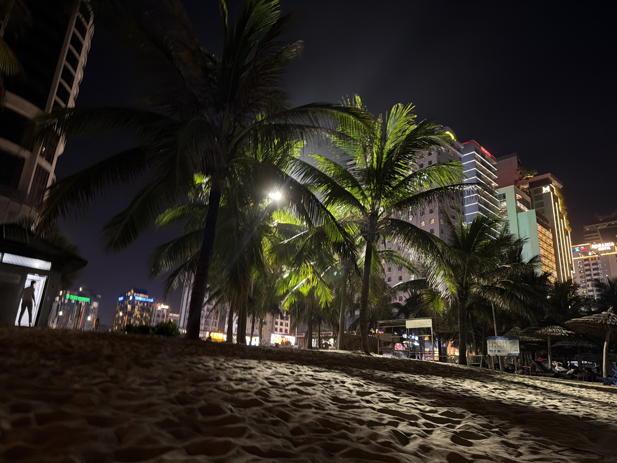 This nighttime image captures a vibrant beach scene in Đà Nẵng, Vietnam, with a focus on artificial lighting. The foreground features textured, golden-brown sand, subtly illuminated to reveal ripples and footprints.
In the mid-ground, numerous tall palm trees stand, their fronds dramatically lit from below by bright artificial lights, giving them a glowing, almost neon-green appearance against the dark sky. To the far left, a silhouetted figure stands within a brightly lit doorway of a small, modern structure. Further into the mid-ground, beach amenities like thatched umbrellas and lounge chairs are visible on the right, suggesting a resort or recreational area. A sign, likely with Vietnamese text detailing beach rules or information, is partially visible near these amenities.
The background is dominated by a modern cityscape of high-rise buildings, many adorned with bright architectural lighting and neon signs that cut through the dark night. Visible text includes "TUAN HOTEL" in red on a building towards the left and "ROSANA" on another building further right. The sky is a clear, dark blue-black, indicating a calm night. The overall impression is one of a lively, illuminated urban beach resort after dusk.
