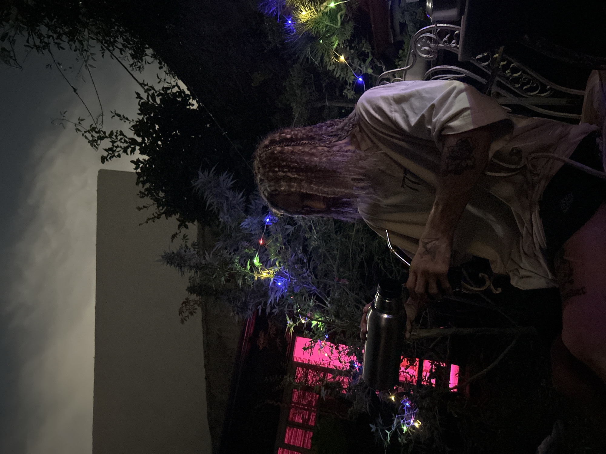A sideways shot shows a person with long, wavy hair, a light-colored shirt, and dark shorts sitting outside at night. The individual is holding a metallic water bottle. Behind the person, there are bushes decorated with colorful string lights. A window with pink lights is visible in the background. A corner of a building is seen on the left, with an overcast sky above. The location cannot be determined from the image.