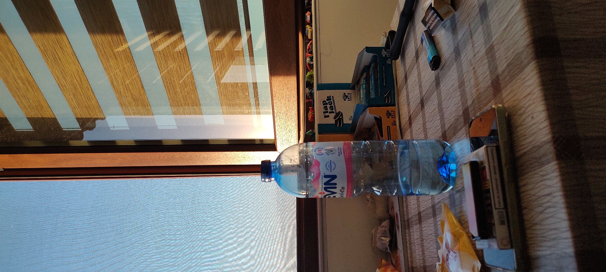 The image shows a room interior with a window on the left side. The window has a brown frame and a partially drawn blind with alternating light and dark brown stripes. A blue screen is visible through the window.
On a surface next to the window, there is a plastic water bottle with a blue cap and a label featuring the word "VIVI". Next to the bottle are several objects stacked, including boxes, what appears to be a lighter, and other miscellaneous items. One of the boxes is labeled "Flap Jack." The surface appears to have a wood-grain pattern.