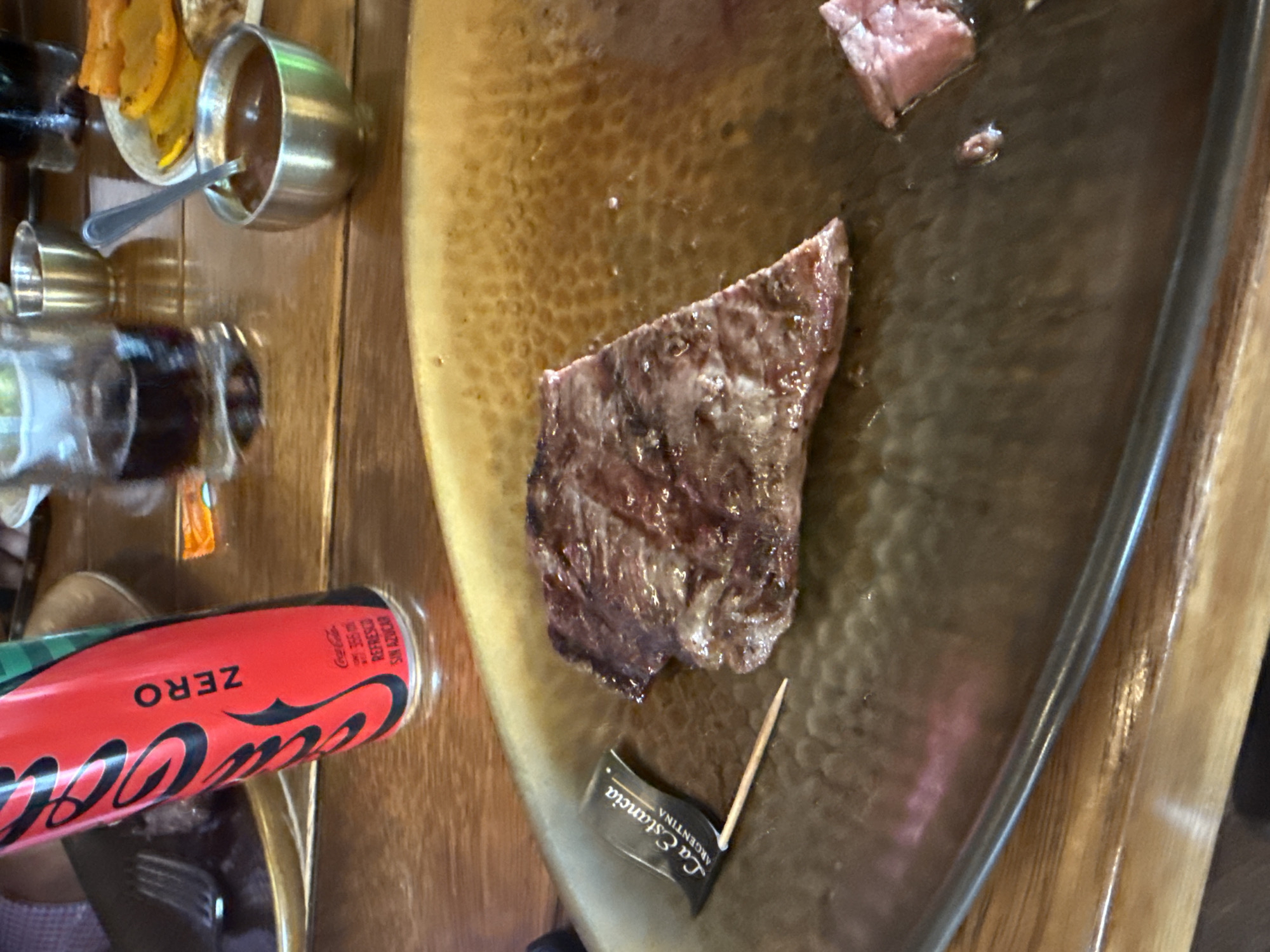 A close-up, high-angle shot captures a meal served on a wooden table, likely within a restaurant setting in San Bernardino Tlaxcalancingo, Mexico. The central focus is a large, textured metal serving platter holding several pieces of cooked steak, indicating an ongoing or recently served meal. One prominent piece of meat, showing a medium-rare doneness, is visible towards the bottom center of the platter.
Accompanying the main dish is a small, polished metal cup with a spoon, containing a dark sauce or condiment, and a small plate holding several slices of golden-brown food, possibly fried plantains or potatoes. A glass bottle or grinder, perhaps for salt or pepper, is also visible on the wooden table.
A red Coca-Cola Zero can rests in the foreground, clearly displaying "ZERO," "Coca-Cola," "REFRESCO SIN AZÚCAR," and Japanese text "ゼロ". A small decorative flag on a toothpick is inserted into the serving platter near the meat, bearing the text "La Estancia" and "ARGENTINA," suggesting an Argentinian restaurant specializing in steak. The overall scene depicts a casual dining experience centered around a meat dish.