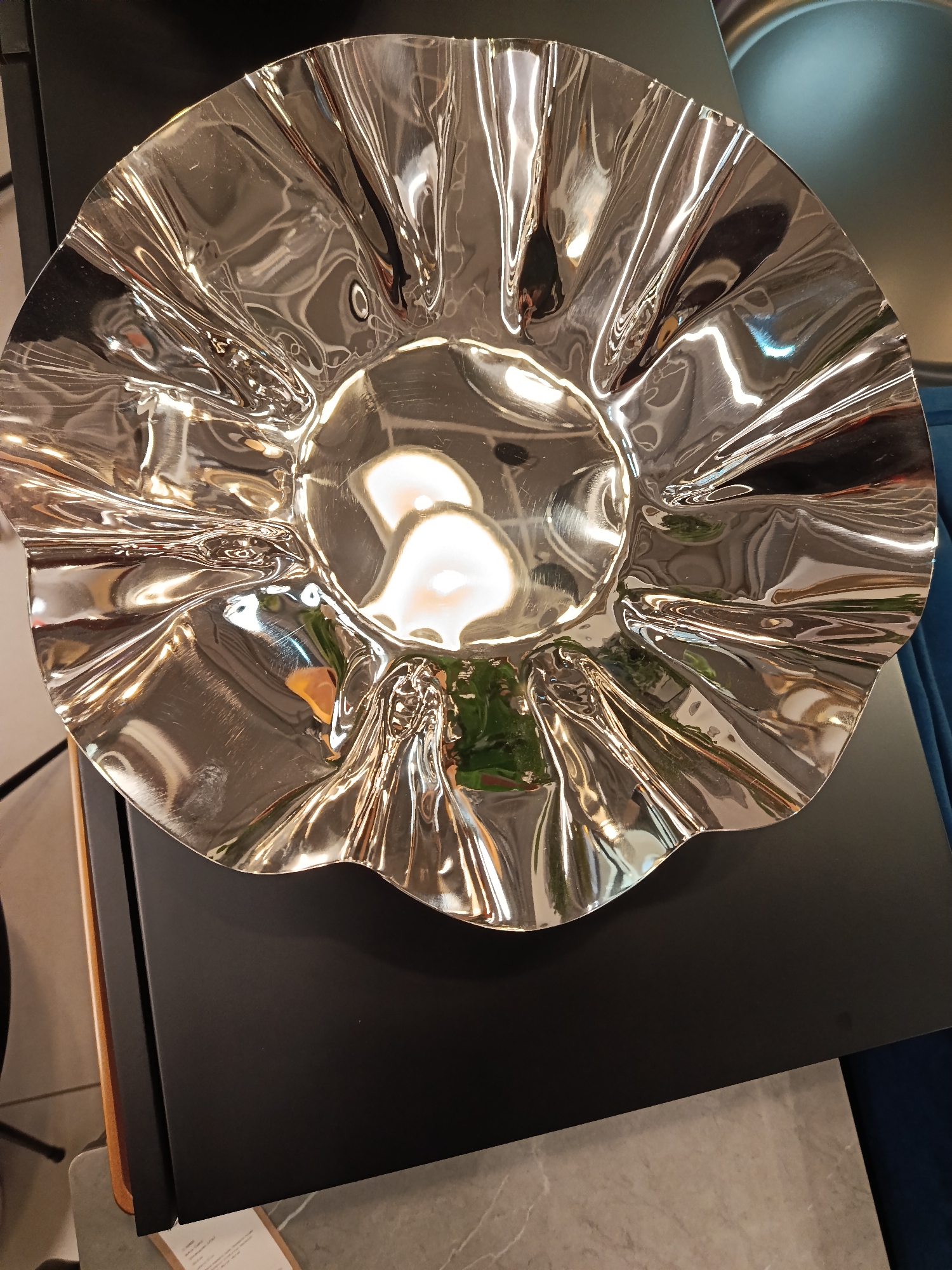 The image displays a large, round, highly reflective metallic decorative bowl or platter with a ruffled, undulating rim and surface. Its polished silver or chrome finish creates significant distortion and reflection of the surrounding environment.
The bowl rests centrally on a flat, dark, rectangular surface, which appears to be a table or display shelf. The highly reflective surface of the bowl captures and distorts bright, elongated indoor light sources, likely overhead fixtures, along with blurred shapes of green foliage (suggesting plants) and other unidentifiable room elements. No people are clearly visible in the image, though indistinct reflections might hint at human forms.
To the left of the dark surface, a partial view of a vertical wooden-toned panel is visible. On the right, a dark blue upholstered surface, possibly part of a chair or sofa, can be seen. The floor beneath the dark surface is a lighter color, appearing to be tiled or marble.
The setting is an indoor environment, likely a contemporary showroom or a modern interior space in Moscow, Russia. The bright artificial lighting suggests the time of day is not a factor. A small, light-colored tag with faint, unreadable text is barely visible on the floor in the lower-left corner. The overall aesthetic is sleek and modern, utilizing reflective surfaces to interact with light and create dynamic visual effects.