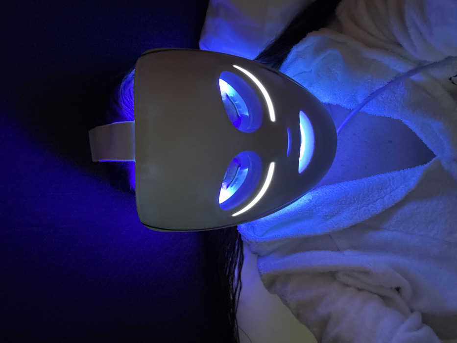 An individual, visible from the chest up, is depicted in an indoor setting in Unknown, Unknown. The person is wearing a white, textured bathrobe and has long, dark hair visible around the sides of their head. Their face is fully covered by a white, rigid facial mask, which emits blue and white light. The mask features two oval-shaped eye openings glowing blue, two curved white light strips mimicking eyebrows, and a curved white light strip mimicking a smile, with a blue glowing mouth opening below. A light-colored strap secures the mask over the top of the head. A thin white cable extends from the lower right side of the mask towards the person's chest. The immediate environment is dimly lit, with the blue light from the mask illuminating the surrounding areas.