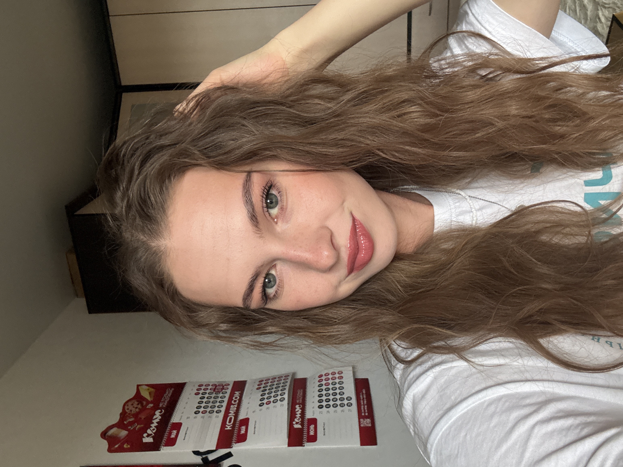 A young female, appearing to be in her late teens or early twenties, is depicted in a close-up, chest-up selfie, captured at eye level in an indoor residential setting in Perm, Russia. She has long, wavy, light brown hair, light blue-green eyes, and a fair complexion with visible freckles across her nose and cheeks. Her lips are painted with a glossy, light pink or nude lipstick, complemented by subtle eye makeup including mascara and light eyeshadow, with faint eyeliner. She is wearing a plain white short-sleeved t-shirt and a thin silver chain necklace. Her left hand is raised to the right side of her head, with fingers gently touching her hair. She presents a slight smile while looking directly at the camera. The background on the left features a red and white wall calendar displaying Cyrillic text, including "КОМУС" (KOMUS) at the top and the months "МАЙ" (May) and "ИЮНЬ" (June). "KOMUS.COM" is also visible at the bottom of the May page. The background on the right shows a section of dark brown furniture, likely a cabinet or wardrobe, against a light-colored wall.