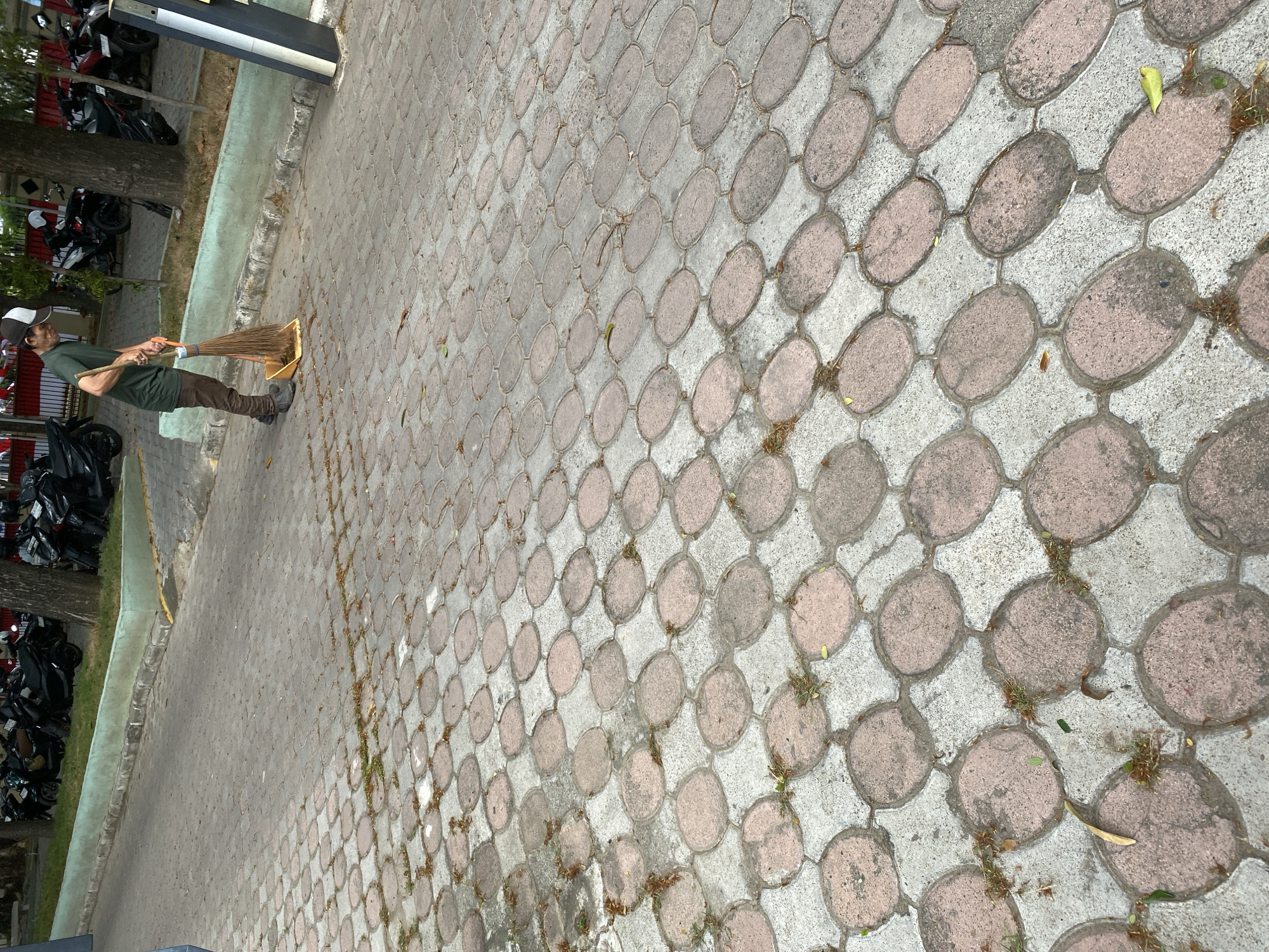 The image depicts an outdoor scene with a man in a green t-shirt and brown pants sweeping a cobblestone pathway with a broom and dustpan. The pathway is paved with alternating oval and rectangular stones, creating a patterned surface. In the background, several motorcycles are parked near trees and a green wall. The environment appears to be a public area within a city, possibly a street or parking lot.