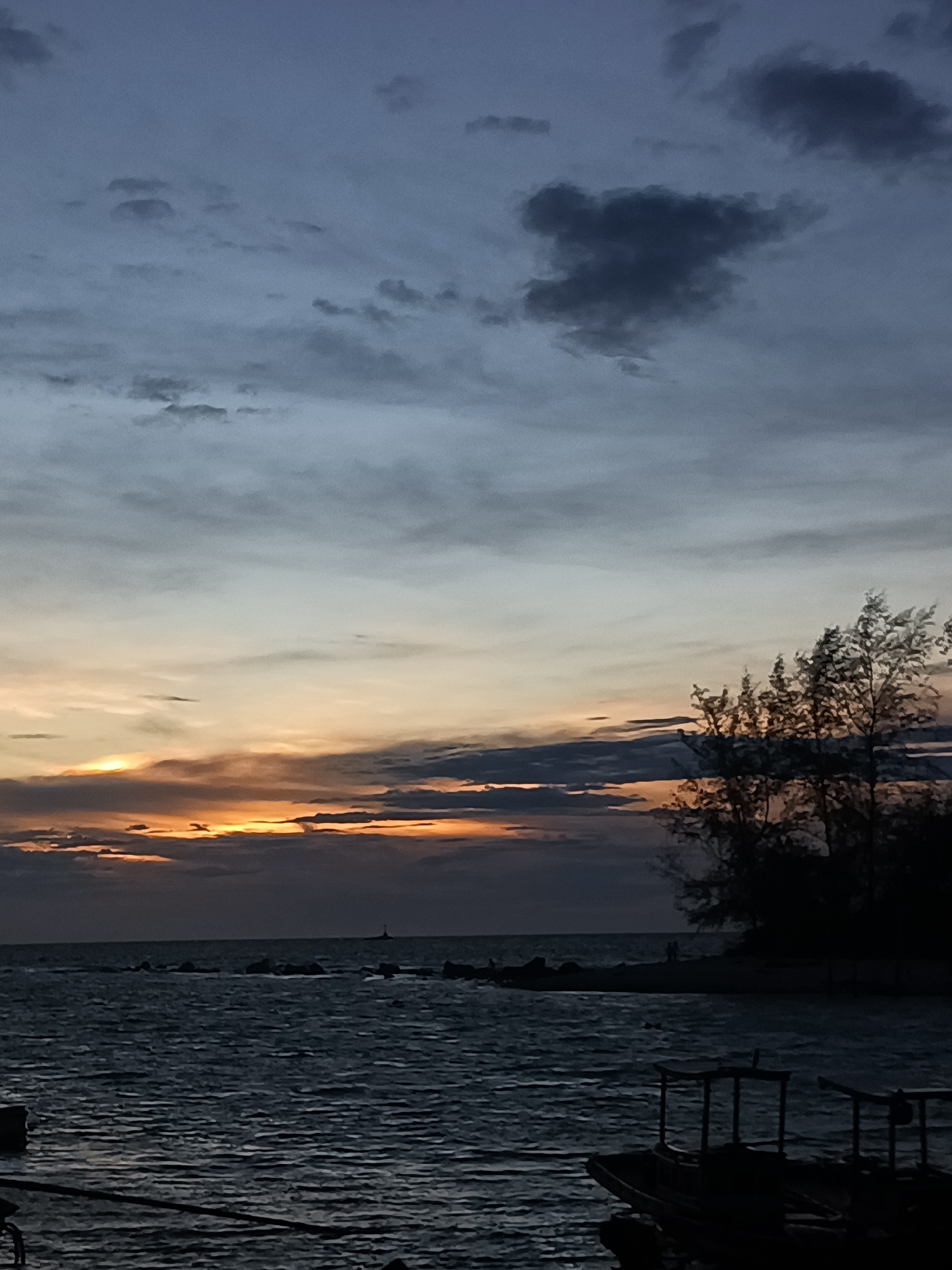 At dusk in Baan Sri Thanu, Thailand, a body of water stretches towards the horizon under a sky with mixed clouds and a band of yellow sunset light. Trees stand silhouetted to the right of the frame. There is a lighthouse or beacon visible in the distance. Boats or structures are visible in the foreground, in silhouette.