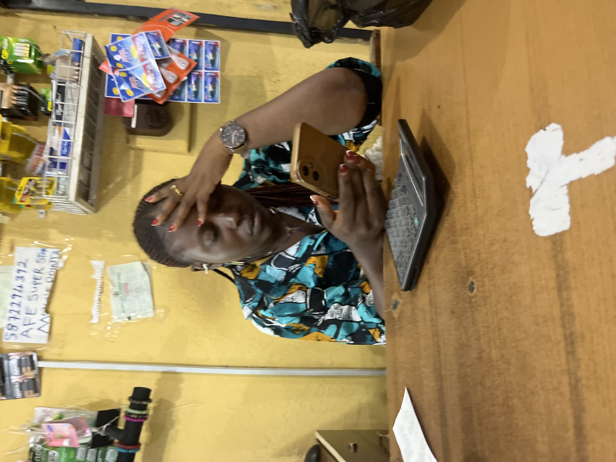 A woman is sitting at a wooden counter in what appears to be a small shop or convenience store. She is wearing a colorful patterned shirt and has a watch on her left wrist. Her right hand is on her forehead, suggesting she might be tired or stressed. She is holding a smartphone in her left hand, and a calculator is on the counter in front of her. Behind her, the wall is painted yellow and displays various items, including packaged goods, batteries, and what looks like a phone number written on a piece of paper. The environment appears somewhat cluttered, with shelves filled with products and some items hanging on the wall. The scene is lit by artificial light. There is no clear indication of interaction with others in the frame. The overall impression is that of a potentially busy or demanding work environment.