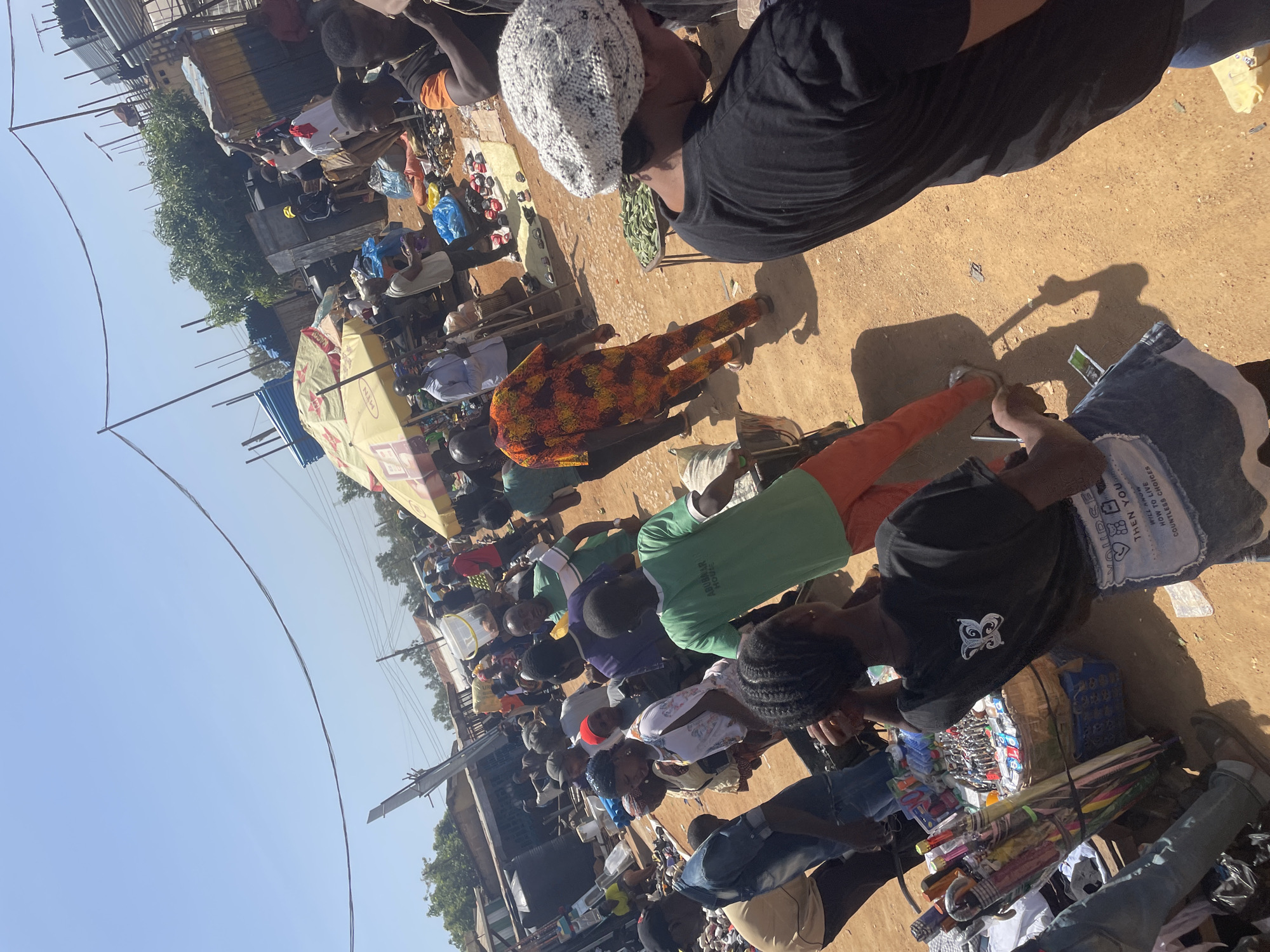 The image portrays a vibrant and bustling outdoor market scene in what is identified as Gura Top, Nigeria. The ground is unpaved, appearing as dry earth or dirt, and the environment is bathed in bright daylight under a clear blue sky, indicated by strong shadows.
Numerous individuals, comprising both vendors and shoppers, are spread throughout the frame. In the foreground, a person wearing a white knitted cap and a black t-shirt looks downwards, while another person in a distinct orange patterned outfit stands further back. Towards the right, an individual with braided hair and a black t-shirt, featuring a white graphic resembling a butterfly or abstract symbol, appears to be attending a market stall. Several people are seen wearing green shirts, one of which clearly displays the text "ABUBAAR HOUSE."
The market itself is characterized by various makeshift stalls, many utilizing large umbrellas for shade. One prominent yellow umbrella bears the "MTN" branding. A wide array of goods is visible, including shoes displayed on the ground, diverse small packaged items, and a bundle of colorful umbrellas for sale at a stall in the lower right. Blue plastic bags are intermittently visible, likely containing purchases. The primary activity depicted is the buying and selling of goods typical of a lively open-air market.
Notable text elements include "MTN" on an umbrella, "ABUBAAR HOUSE" on a green shirt, and a fragmented phrase on denim clothing or a bag in the lower right, reading "THEN YOU WILL ANOW HOW TO LIVE COUNTLESS CHOICES." The overall impression is one of everyday commerce and community activity under a sunny sky.