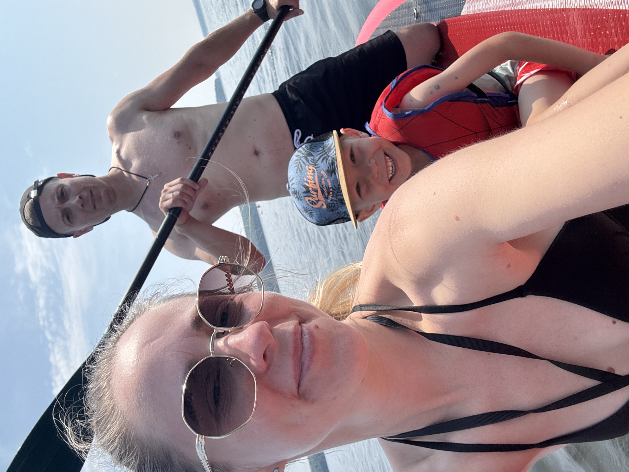 This is a portrait-oriented, outdoor, selfie-style photograph taken on a paddleboard in Chaikovsky, Russia. Three people are visible: a man and a woman in the foreground, and a young boy in the middle ground. The subjects are on a red paddleboard on calm water, under a bright, partly cloudy sky, suggesting a sunny day.
The woman in the foreground is wearing sunglasses and a black swimsuit with intricate straps. She is looking directly at the camera with a relaxed expression. The man, positioned behind her and to the left, is shirtless and wearing black shorts and a black cap. He is holding a paddle, with part of it visible. The young boy is in the middle ground, wearing a red and blue life vest and a blue baseball cap with a palm tree design and text. He is smiling broadly at the camera, and appears to be lying down or leaning back on the board.
The text on the boy's cap reads "Surfing" and "Coastal Vibes". The overall mood of the photograph is joyful and suggests a fun outdoor activity. The lighting and the presence of water indicate it is daytime, likely late morning or afternoon.