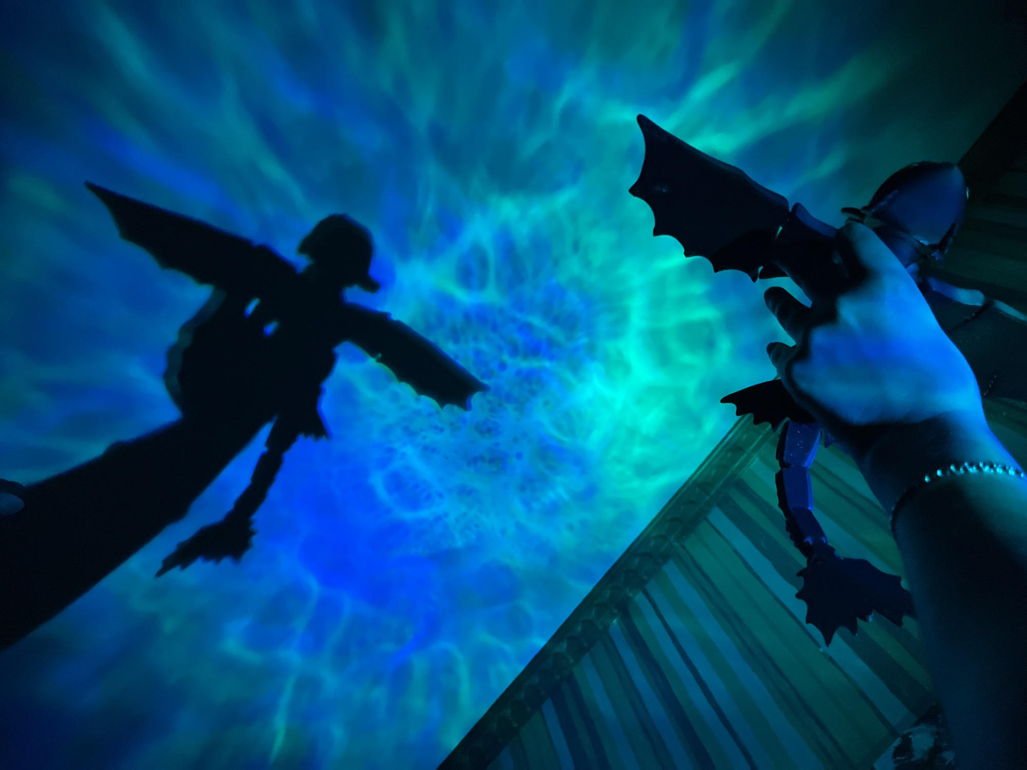 A person's hand holds a black silhouette of a dragon against a backdrop of blue and green swirling light patterns that resemble water or aurora borealis. Above, a larger, similar silhouette of a dragon is projected onto the same illuminated surface. To the lower left, a striped surface is visible, with diagonal lines in shades of blue and green. The scene appears to be indoors, with the lighting creating a dramatic, otherworldly effect. The specific location in Saint Petersburg, Russia, cannot be determined from the visual information provided.