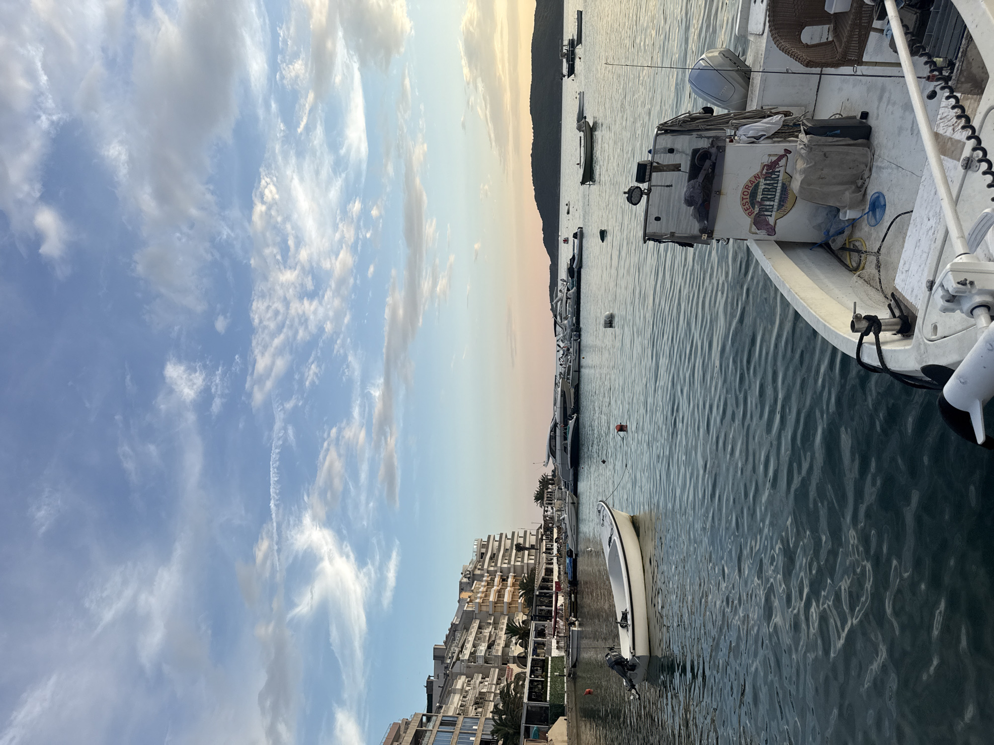 This is a vertical, eye-level, medium shot of a marina in Rafailovići, Montenegro, likely taken during the golden hour, as suggested by the warm tones in the sky and the soft light. The sky is a prominent feature, filled with dynamic, wispy clouds against a gradient of blue and soft pink. Below the sky, a dark, mountainous silhouette is visible in the distance.
The foreground is dominated by a body of water, reflecting the sky and creating rippling patterns. Several boats are moored in the marina. The closest boat, on the right side of the frame, is a white fishing vessel with an outboard motor at its stern and various fishing equipment, including fishing rods and a net. A logo on the rear of this boat reads "RESTORAN ARUBA." A woven chair is also visible on this boat.
To the left of the water, a dense row of multi-story buildings lines the waterfront. These buildings have a consistent architectural style with numerous balconies and windows, suggesting a busy tourist or residential area. Palm trees are interspersed among the buildings, adding to the Mediterranean atmosphere.
The scene appears peaceful and serene, with no visible people. The calm water and the soft lighting contribute to a tranquil ambiance. The location cues, such as the architecture and the presence of boats, firmly place the scene in a coastal European setting, specifically Rafailovići, Montenegro.