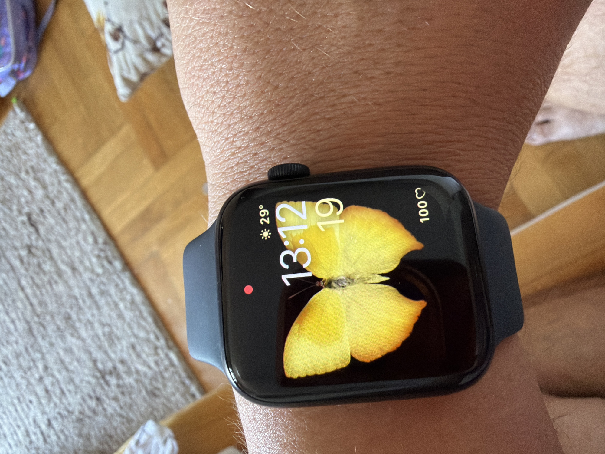 The image shows a close-up of a person's wrist wearing a smartwatch. The smartwatch has a black band and a dark screen displaying a vibrant yellow butterfly as the watch face. The time on the watch reads 13:12, and it is the 19th of the month. To the left of the time, the temperature is displayed as 29° with a sun icon, indicating sunny weather. On the right side of the screen, a heart icon is shown next to the number 100, likely representing a heart rate or activity metric.
The background is slightly out of focus, revealing hints of an indoor setting with wooden flooring and a textured rug. There are also blurred elements that suggest soft furnishings, possibly on a bed or sofa. The lighting appears natural, suggesting daytime. The overall mood conveyed is calm and everyday, with the focus on the technology and the visually appealing watch face. There are no explicit human figures visible beyond the wrist. The location context is Budva, Montenegro, though this is not directly discernible from the visual elements in the image itself.