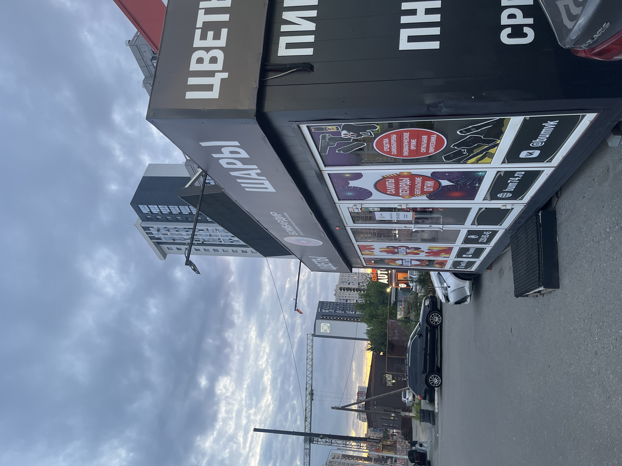 The image depicts a commercial storefront in Chelyabinsk, Russia. The store appears to sell a variety of items including balloons ("ШАРЫ"), flowers ("ЦВЕТН"), and pyrotechnics such as fireworks ("САЛЮТЫ", "ПЕТАРДЫ", "БЕНГАЛЬСКИЕ ОГНИ") and signal flares ("СИГНАЛЬНАЯ ПИРОТЕХНИКА"). There are also advertisements for self-defense items ("СРЕДСТВА САМООБОРОНЫ") and pneumatic weapons ("ПНЕВМАТИЧЕСКОЕ ОРУЖИЕ").
Visible text includes operating hours "10:00-20:00", a phone number "220-85-45", a website "bum74.ru", and social media handles "@bumvk". The name "AUT" is also visible, possibly referring to an automotive section or service. The word "SOLARIS" is seen on the taillight of a car, indicating the make of the vehicle.
The scene is set outdoors, with a cloudy sky suggesting it might be late afternoon or early evening. The sky has a mix of dark and lighter clouds, with a hint of sunset colors visible in the distance behind buildings. Several buildings, including a tall apartment complex, are visible in the background, along with a construction crane, suggesting ongoing development in the area. Two cars are parked in front of the store. The overall impression is that of a commercial street scene under a dynamic sky. No people are clearly visible in the image.