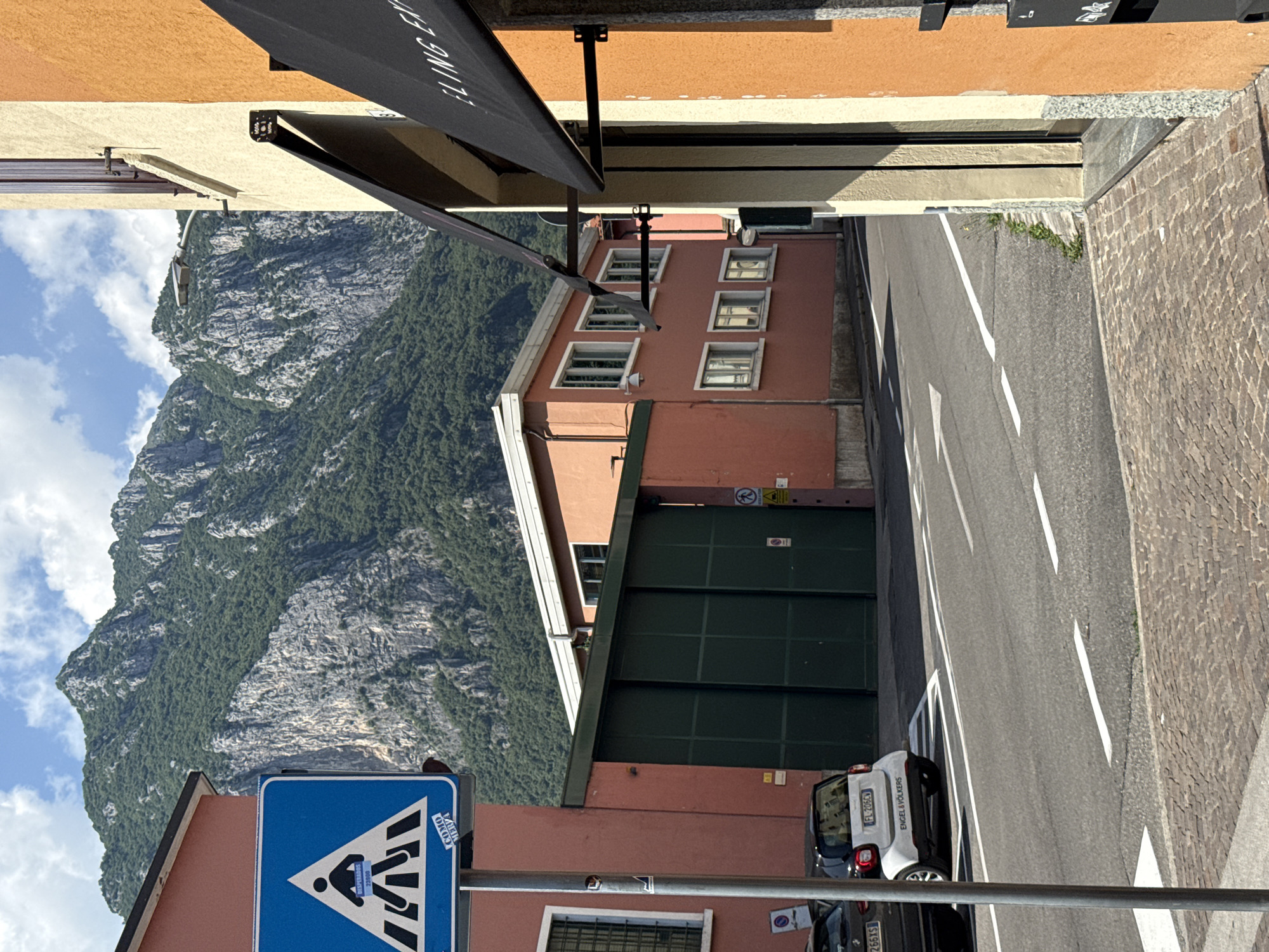 A street scene is depicted, with buildings lining one side and a steep, tree-covered mountain in the background under a bright, partially cloudy sky. In the foreground, a pedestrian crossing sign is visible, a blue square with a white triangle containing a silhouette of people crossing a street. A small white car is parked near the curb. The street is paved with asphalt and marked with white lines, indicating traffic lanes and a pedestrian crossing. On the right side of the image, a building with an orange facade and a black awning is prominent.