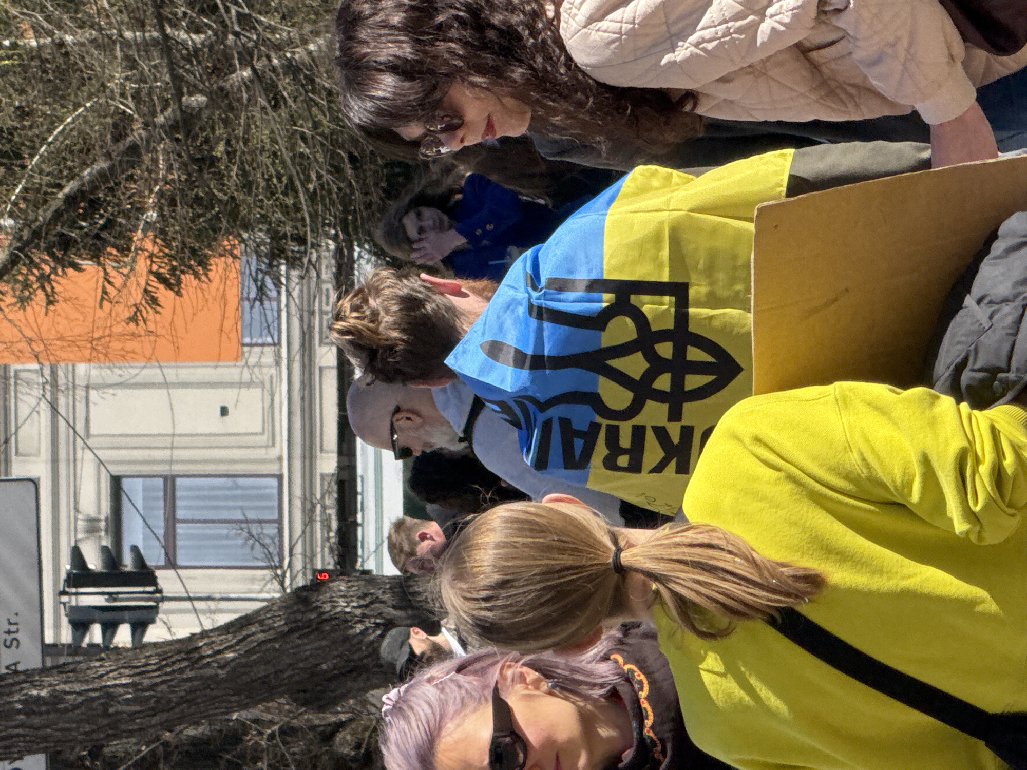 The image shows a daytime street scene in what appears to be a Western European city. A group of people is gathered, some holding a flag displaying the colors of the Ukrainian flag (blue and yellow) and the Ukrainian trident symbol. The word "UKRAINE" is also visible on the flag, oriented upside-down in the image's perspective. People in the group are dressed in various casual clothes, including a yellow sweatshirt, a quilted jacket, and a blue jacket. A portion of a building is visible in the background, featuring an orange facade and windows. There is also a tree branch and a street sign. Without more information, it is not possible to determine whether the location is Kyiv, Ukraine. The presence of the Ukrainian flag suggests a connection to Ukraine, but it does not definitively confirm the location.
