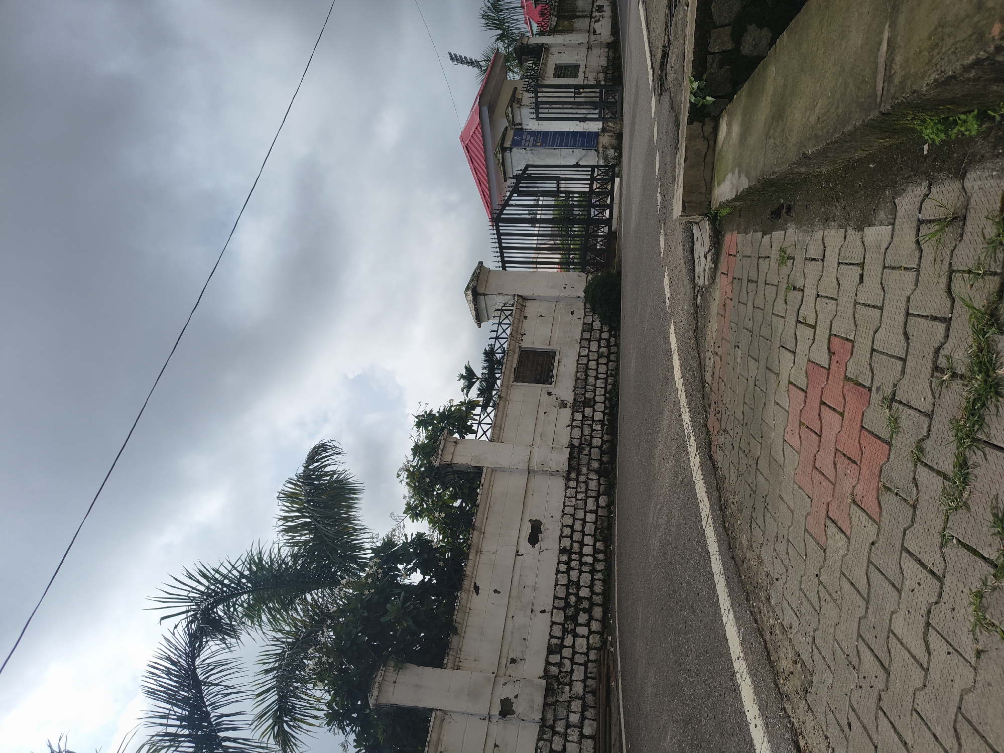 This image shows a street view in Dharamshala, India, featuring a white wall with a stone foundation, topped with trees and foliage. Beyond the wall, a black metal gate leads to a building with a red roof. The street in the foreground is paved with grey and red interlocking bricks, with a dark asphalt road to the left, marked by a white line. The sky is overcast with grey clouds, and a power line stretches diagonally across the top of the frame. No people are visible in the image.