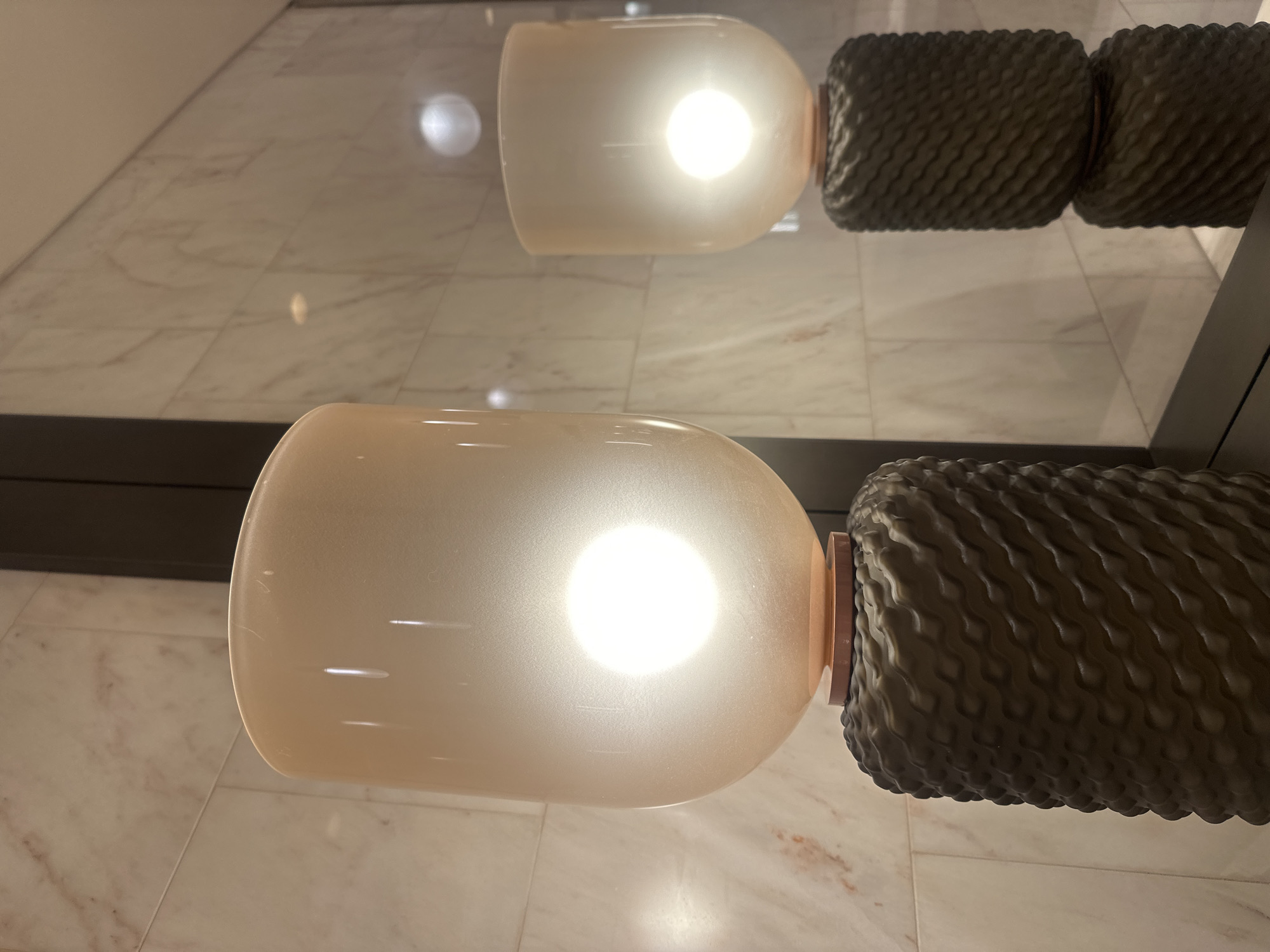 The image is a close-up photograph of a modern light fixture in what appears to be a contemporary interior. The fixture consists of a rounded, elongated glass shade that glows with a warm light emanating from within. This shade is attached to a textured, cylindrical component made of dark material with a repetitive, possibly woven pattern. The texture is reminiscent of a coiled rope or a basket weave. The reflection of the same fixture is faintly visible in the background. The surface below is a light-colored, possibly marble or tiled floor with subtle veining patterns. The presence of a dark line in the background suggests a door or panel. There are no people visible in the photograph. The location in the city of Milan, Italy is not discernible based solely on the image.