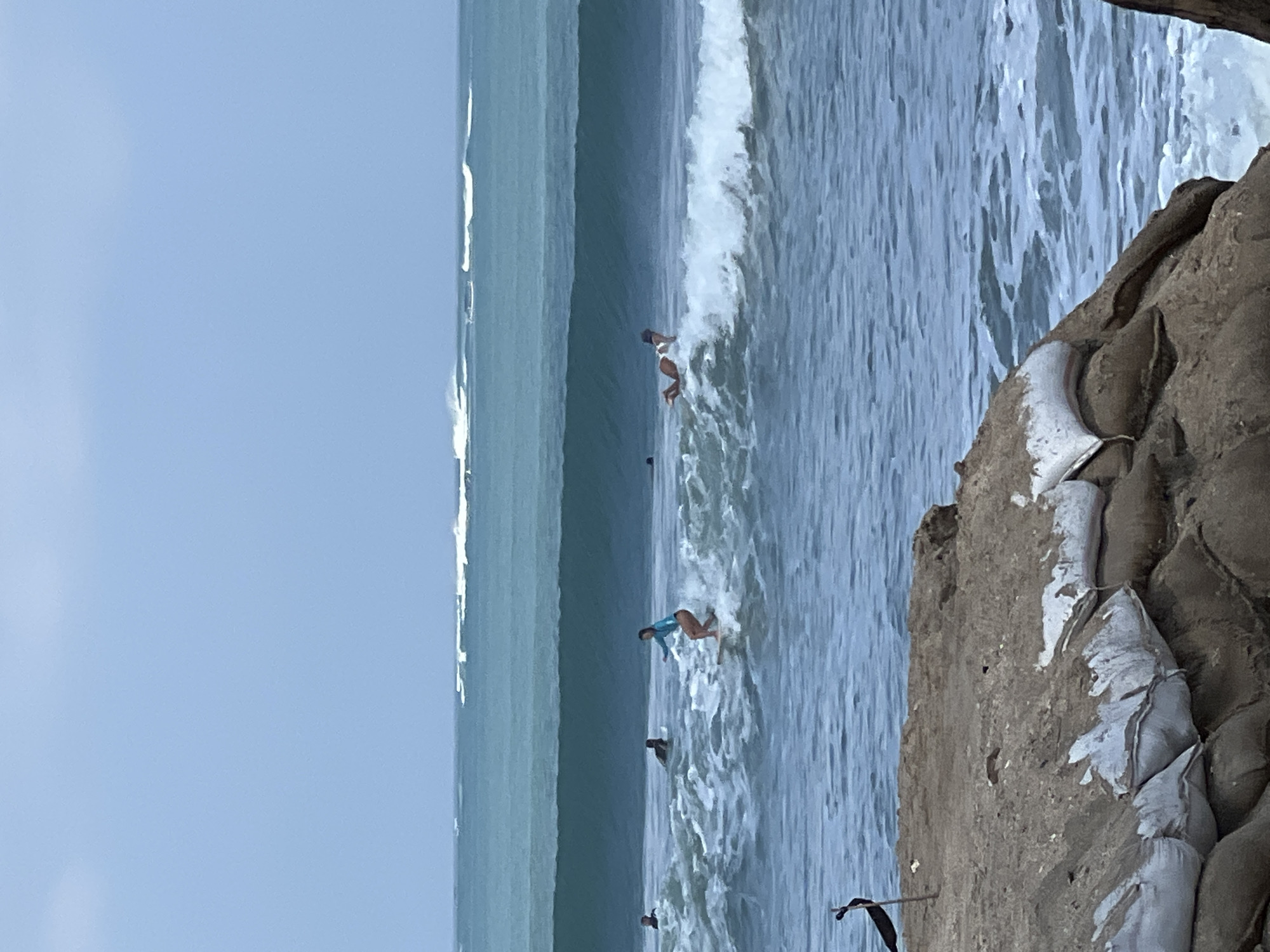 In Kuta, Indonesia, this image captures surfers navigating the ocean. The foreground features a sandy embankment reinforced with sandbags, some of which are torn and exposed. A dark, thin pole with a black flag-like object is planted in the sand to the left. The middle ground shows several individuals on surfboards in the water. One surfer in a blue top and brown bottoms is actively riding a wave, kicking up white spray. Further out, another surfer in a white bikini is also on a wave. More surfers are visible in the distance, some paddling. The background is dominated by the ocean with multiple lines of waves breaking, and a hazy, pale blue sky above. The overall scene depicts a typical day at a surfing beach.