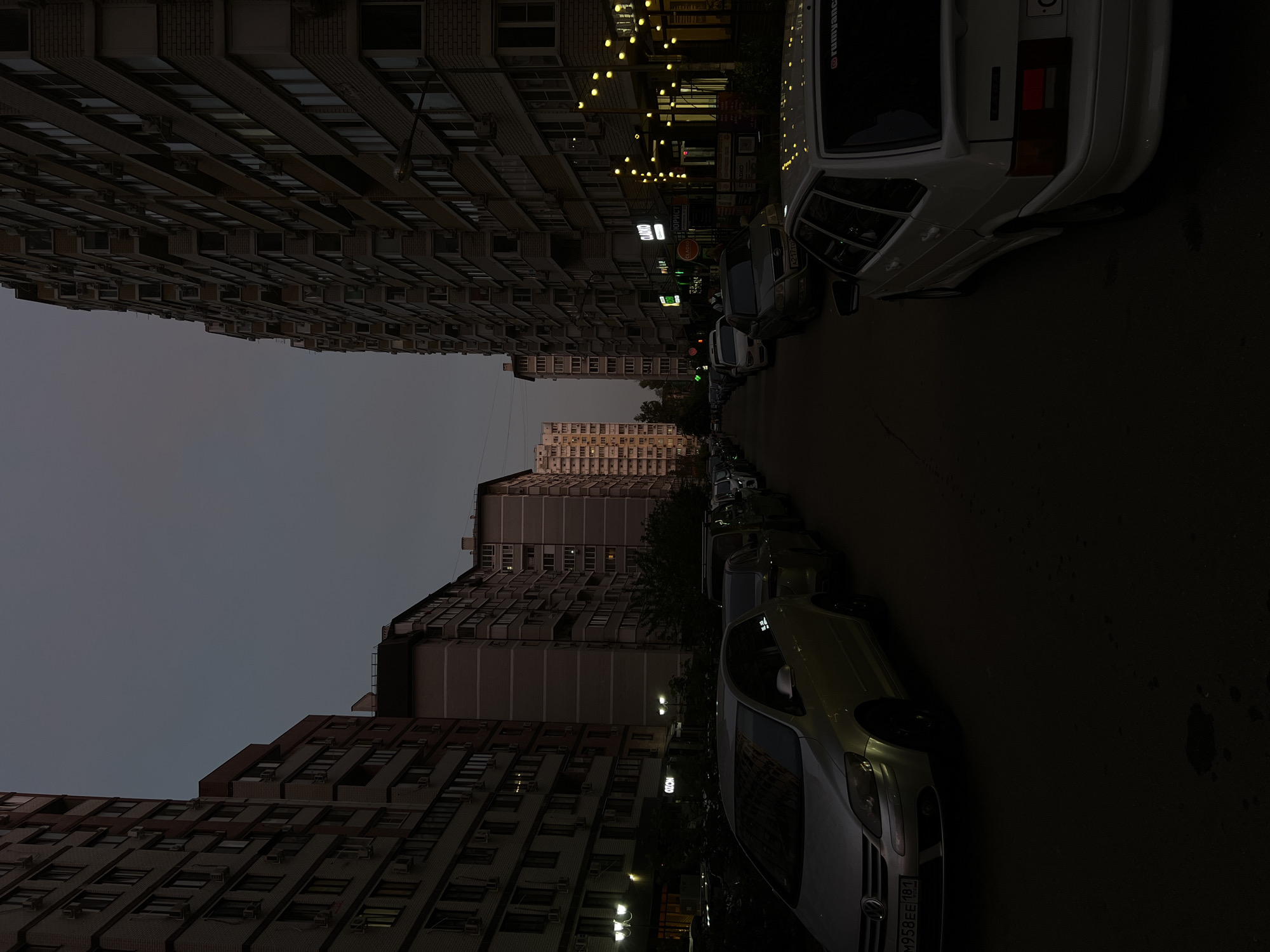 Here is a forensic description of the image:
The image captures an urban street scene in what appears to be the city of Krasnodar, Russia, based on the Cyrillic script visible on a car license plate. Tall residential buildings line both sides of the street. Several parked vehicles occupy the street's edge, including a light-colored car with a recognizable license plate reading "М958ЕЕ 181." A building in the distance is illuminated by the setting sun. The general lighting suggests that it is dusk or a dimly lit environment.