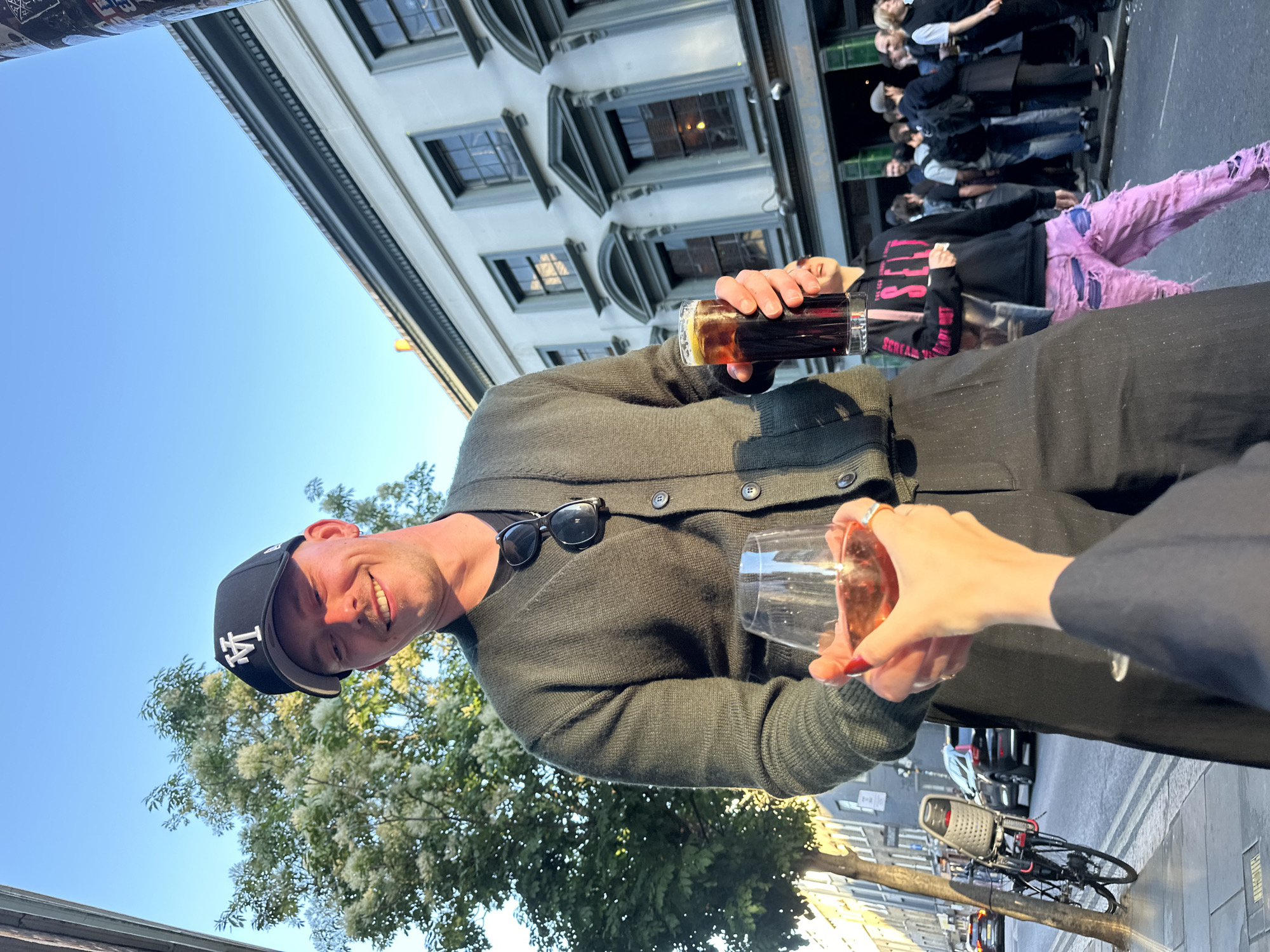 The image, captured from a low angle and rotated 90 degrees clockwise, presents a vibrant outdoor social scene in an urban setting, likely London.
The main subject, a smiling man, occupies the left side of the frame. He wears a dark green knitted cardigan and a black baseball cap with a white "LA" logo. Black sunglasses are tucked into his cardigan. He holds a tall glass containing a dark beverage (possibly a stout or dark mixed drink) and is clinking it with a wine glass held by another person's hand, which is adorned with red nail polish and a ring. This wine glass contains a pinkish drink, likely rosé or a cocktail.
In the background, an urban street scene unfolds. A group of people is visible, some appearing to be queuing outside a building. One individual stands out wearing a black hoodie with partially visible pink text, "SCREAM," and distinctly ripped, light pink trousers. Further down the street, a black bicycle with a front basket is parked on the sidewalk.
The background features multi-storied buildings with classic European-style architecture, including ornate window frames and cornices, suggesting a historical urban environment. The sky is a clear, bright blue, indicating good weather. Lush green trees with small white blossoms are visible in the lower left portion of the image.
The warm, bright light suggests late afternoon or early evening on a sunny day. The man's smile and the act of clinking glasses convey a friendly, celebratory, and joyful atmosphere.
