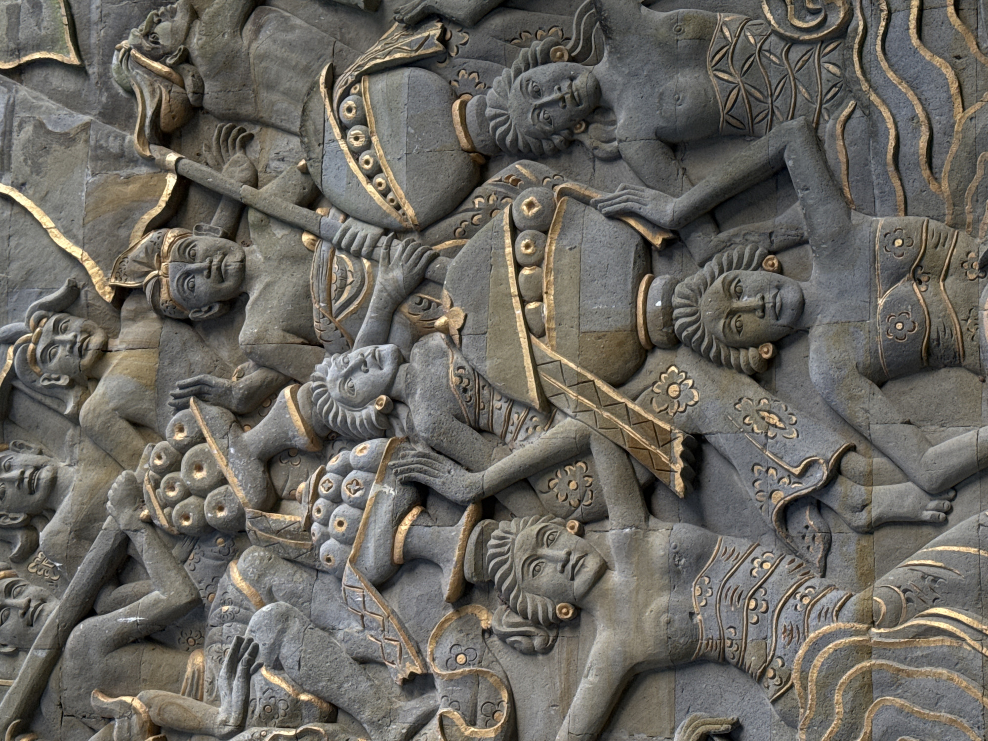 This is a close-up image of a gray stone bas-relief with golden-colored highlights, located in Kuta Selatan, Indonesia. The relief depicts several human figures lying horizontally, many seemingly overlapping or intertwined. Some figures are adorned with ornate garments or armor-like coverings, decorated with golden embellishments. One figure holds a long object that resembles a spear or staff. The overall composition suggests a scene of conflict or chaos. The carvings have intricate details, with patterned robes and other ornamental designs. The style of the relief suggests a Balinese or Southeast Asian artistic tradition.