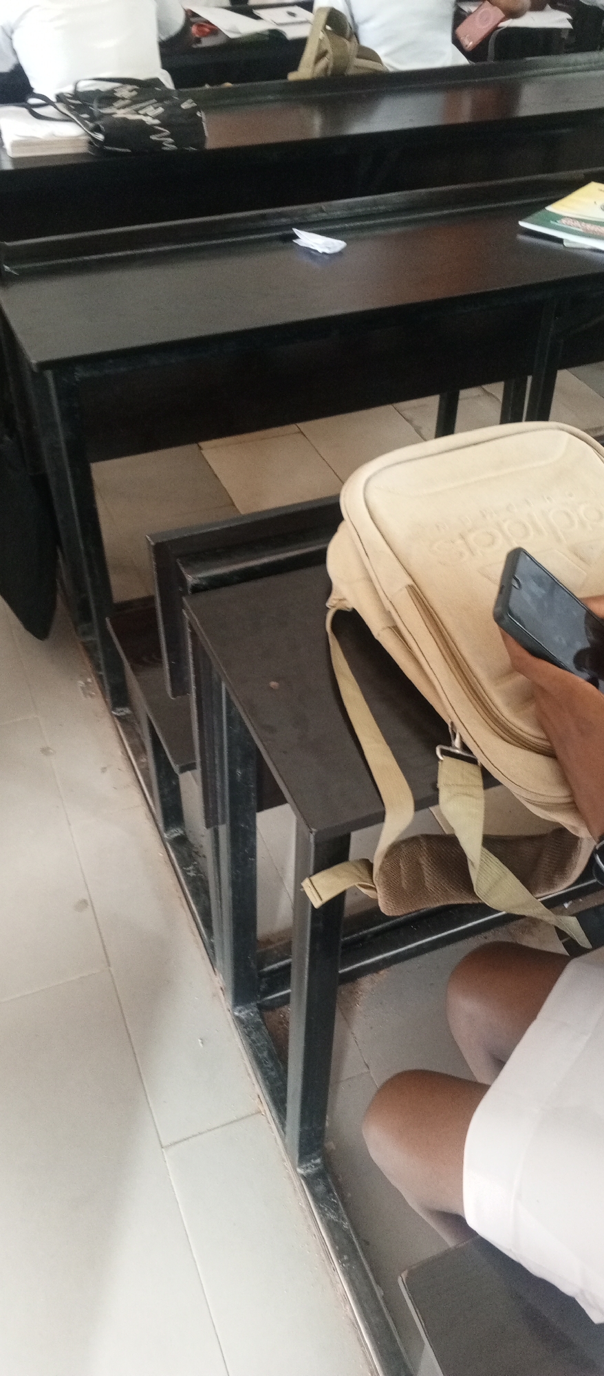 The image captures an indoor scene, likely a classroom or lecture hall, with rows of dark, tiered desks. The focus is on the lower half of the frame, showing a beige crossbody bag with an embossed logo resting on a desk. A person's hand is visible holding a smartphone, and their legs, clad in white, are also in view, suggesting they are seated. The floor is tiled in a light beige color.
In the background, other desks are visible, some with objects on them, including what appears to be a black patterned bag, white paper, and a book. The overall impression is of an educational setting, possibly during a class or break. There are no strong indicators of the time of day or weather conditions, but the indoor lighting suggests it's daytime.
The embossed logo on the bag appears to be "adidas." The smartphone in hand is dark-colored. The scene is set in Kuson, Nigeria. The mood is neutral, depicting a common everyday situation in an academic environment.