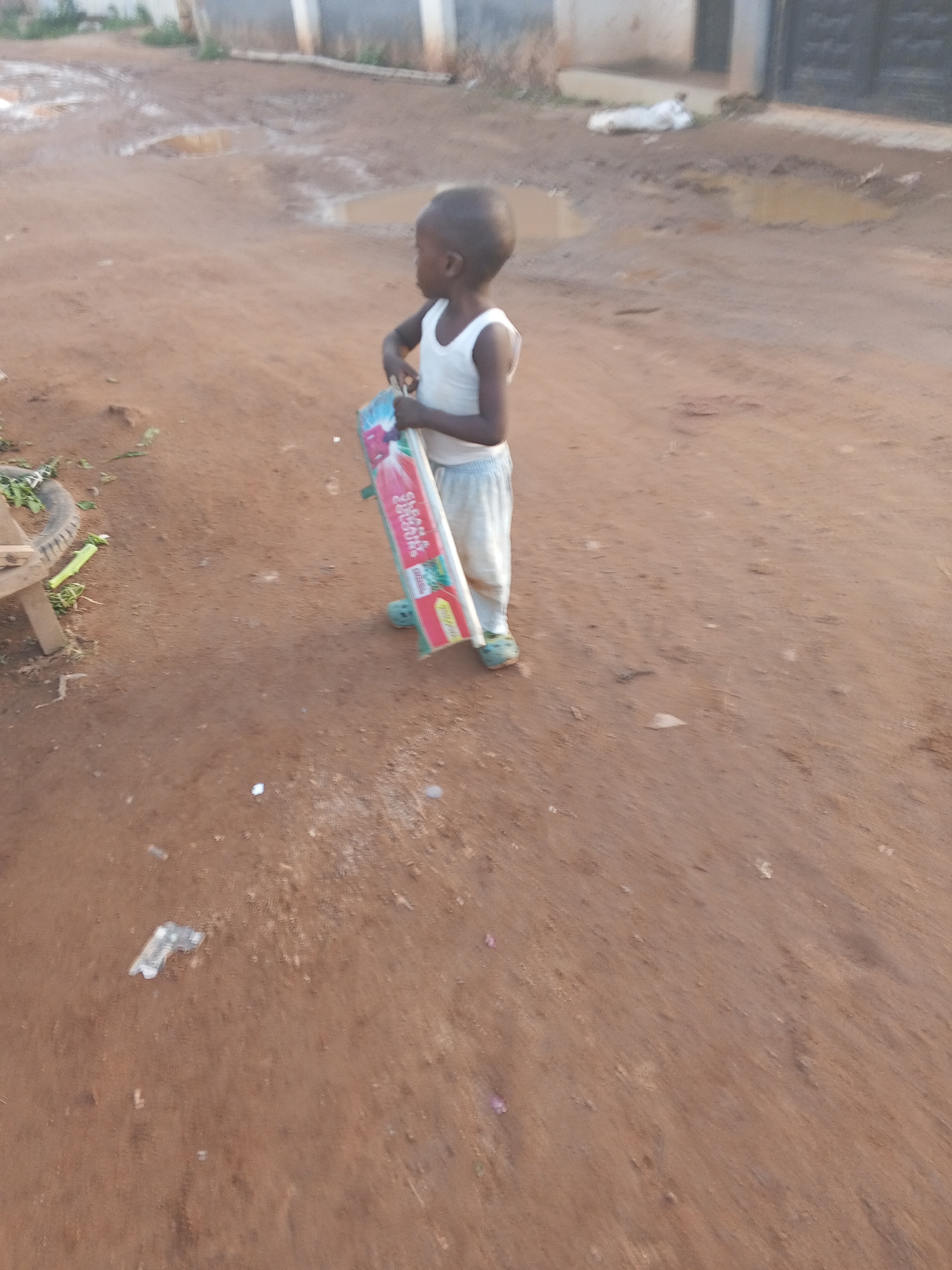 A young Black child in a white tank top and light-colored pants stands on a dirt road. The child holds a long, rectangular package with pink and red colors. Puddles are visible on the road behind the child. A tire rests beside a crude wooden stool on the left side of the image. In the background, a partially visible building with grey and white elements and a closed black gate can be seen. The location appears to be a residential or peri-urban area in Abuja, Nigeria.