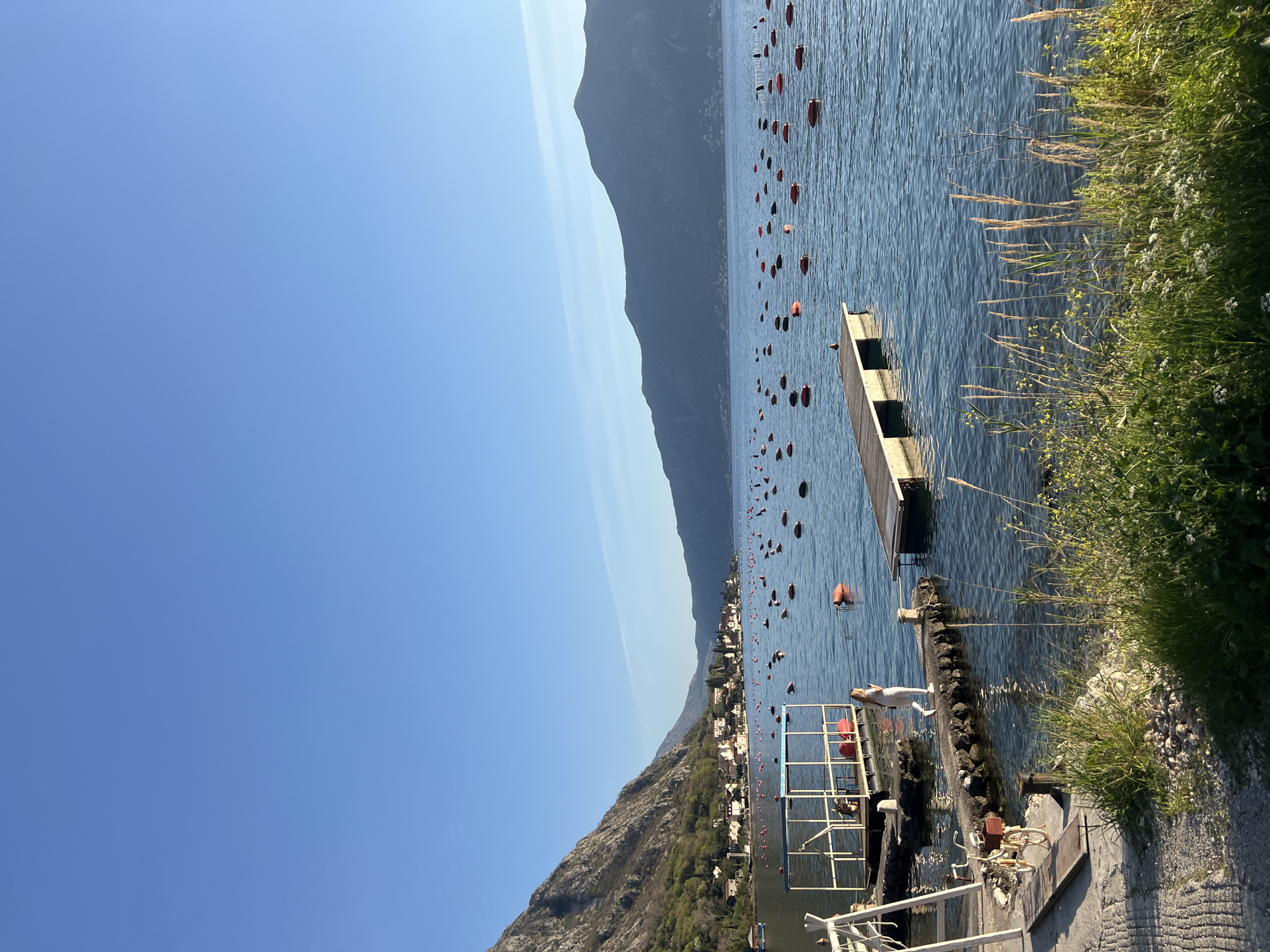 The image captures a sunny day at the Bay of Kotor in Donji Orahovac, Montenegro. A portion of a pier with a metal railing extends into the water. A woman in a white dress stands on the pier. Numerous orange and red buoys are scattered across the bay. In the background, a mountain range is visible, with a town nestled along its base. Vegetation grows along the edge of the pier.