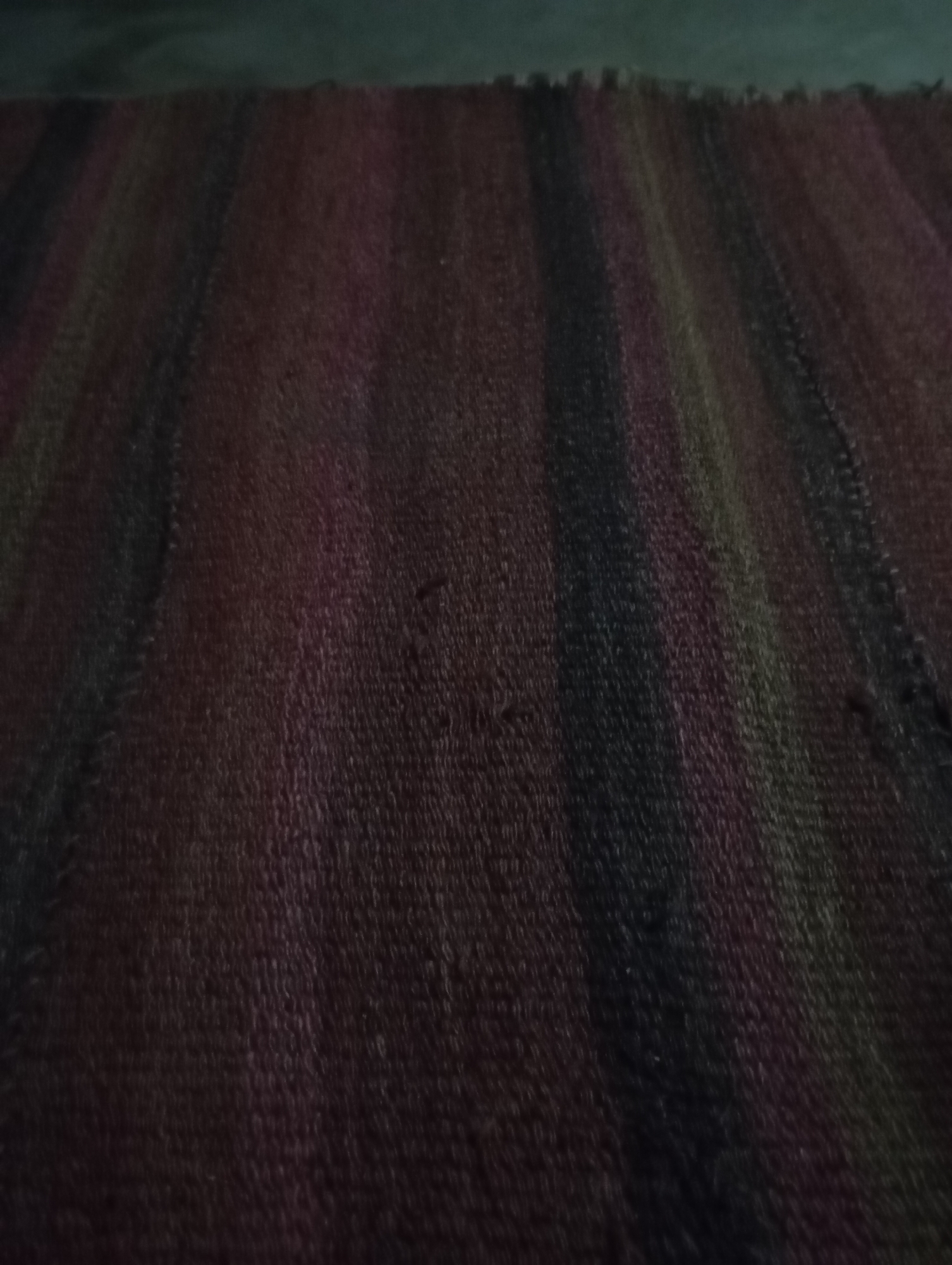 The image shows a close-up, overhead view of a textured rug with vertical stripes. The rug appears to be made of woven fabric, with visible thread patterns creating a raised surface. The colors of the stripes include shades of dark red, purple, black, and hints of green and yellow. The lighting is dim, casting shadows and making some colors appear darker than they might be in brighter conditions. The rug is laid out on a dark, likely floor surface, visible at the top edge of the image. There are no people or discernible objects other than the rug itself. The scene is static and offers no indication of activity or a specific location within a city like Samarqand, Uzbekistan.