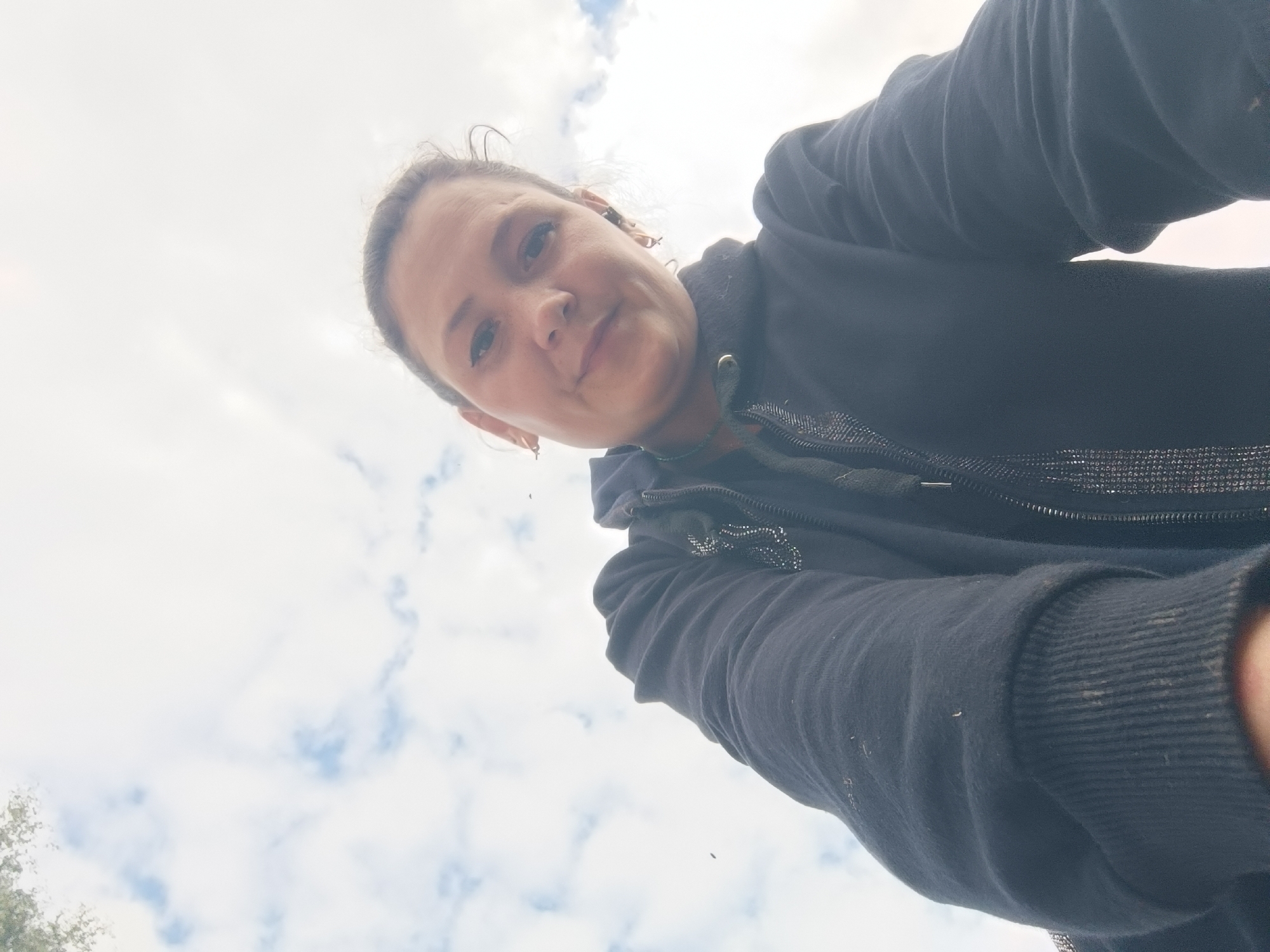 A low-angle, over-the-shoulder selfie is taken by a woman with dark hair pulled back, looking directly at the camera. She is wearing a dark zip-up hooded jacket with some subtle sparkle detail around the zipper. The background is dominated by a pale sky with scattered white and light blue clouds. The image is slightly overexposed, suggesting bright outdoor lighting. There are no discernible urban features or landmarks that would place the location specifically in the city of Karsha, Russia. The scene appears to be an outdoor environment, possibly a park or open area, with a hint of foliage visible in the lower left corner.
