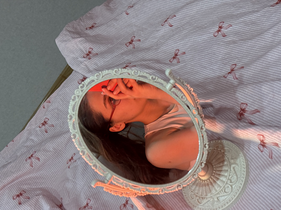 The image depicts a mirror selfie taken inside a private residence, specifically a bedroom, in Krasnodar, Russia. An ornate, oval-shaped, cream-colored vanity mirror with a decorative base is positioned on a bed. The bed is covered with light pink and white striped bedsheets featuring a repeating pattern of small red bows. Reflected in the mirror is a female subject with long, dark hair, wearing red-rimmed glasses and a light-colored tank top or camisole. Her right hand is visible, holding a bright red smartphone, which partially obscures her face, specifically the nose and mouth area, indicating the act of taking the photograph. Her fingernails are painted a light color. The entire scene is bathed in a distinct warm, orange-red ambient light.
